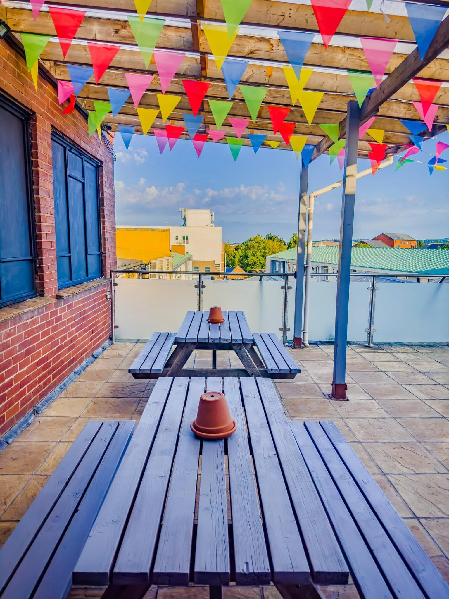 Rooftop terrace with wooden picnic tables, orange flowerpots, and colorful triangular pennant banners hanging from a wooden pergola, with city buildings and partly cloudy sky in the background.