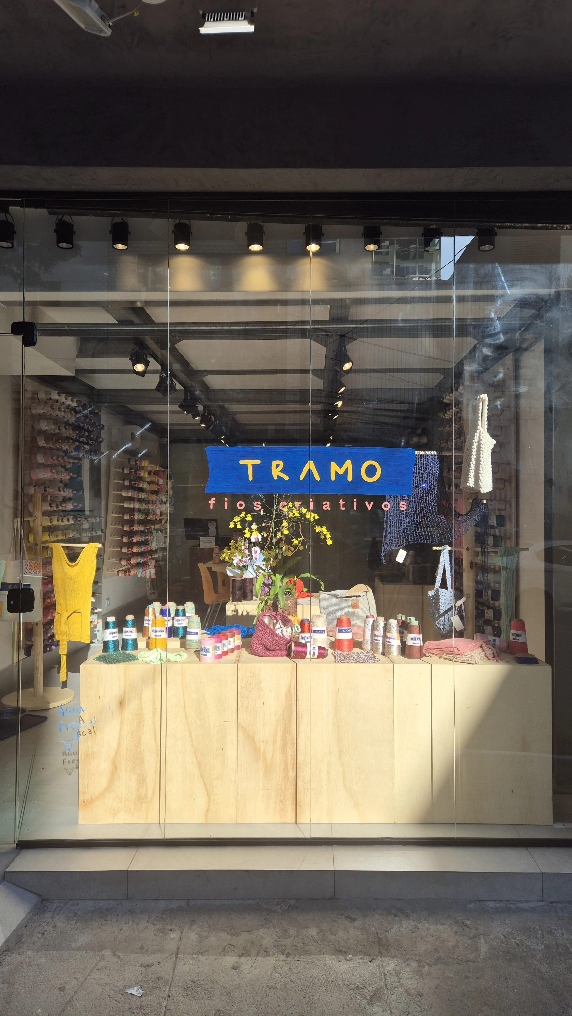 Vitrine de loja que exibe produtos de crochê e tricô, com o nome 'TRAMO fios criativos' na vitrine, decorada com plantas e objetos de artesanato.