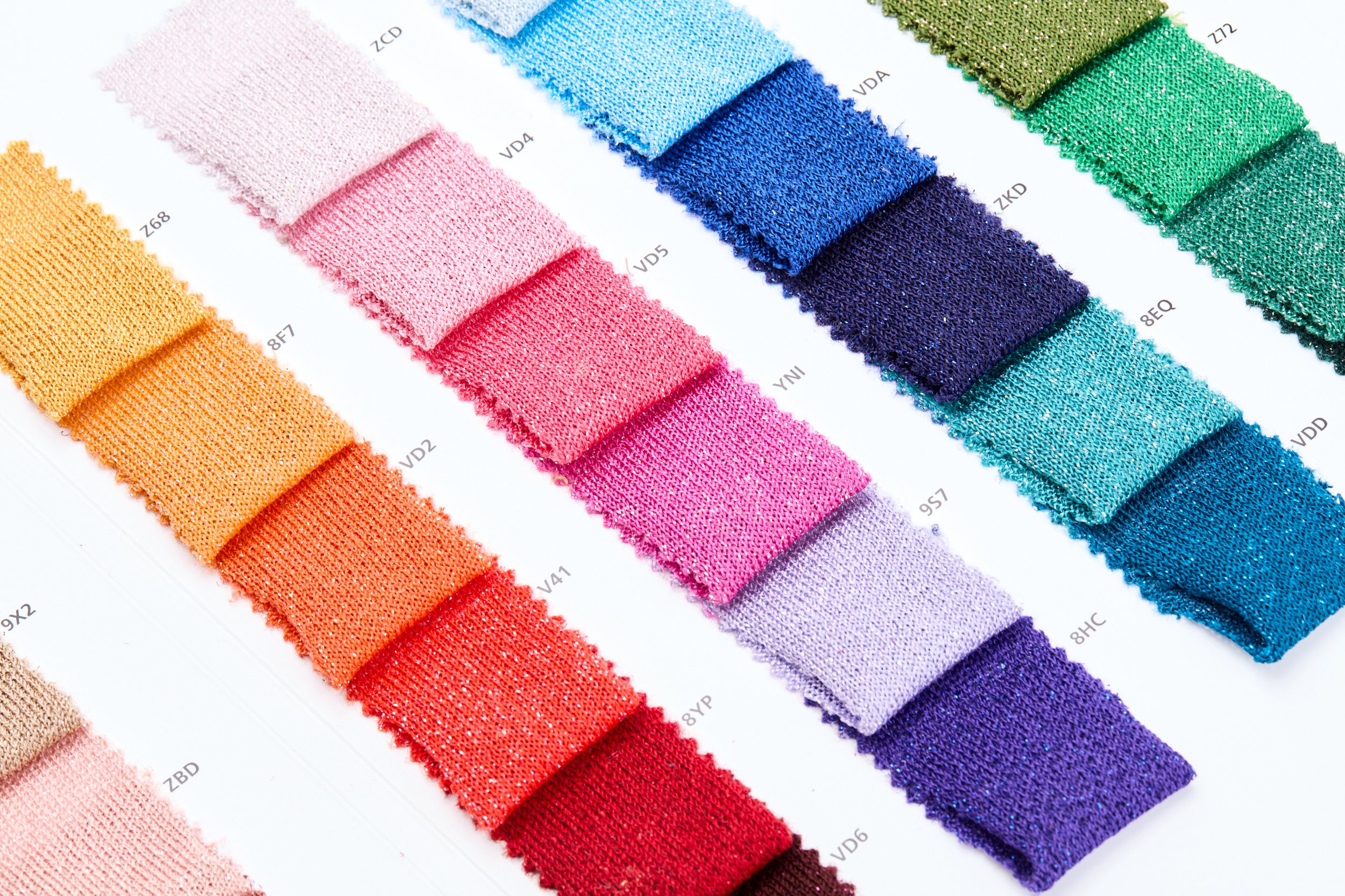 Mostruário de tiras de tecido coloridas dispostas em linhas, mostrando várias cores e texturas, cada uma com uma etiqueta de código ao lado.