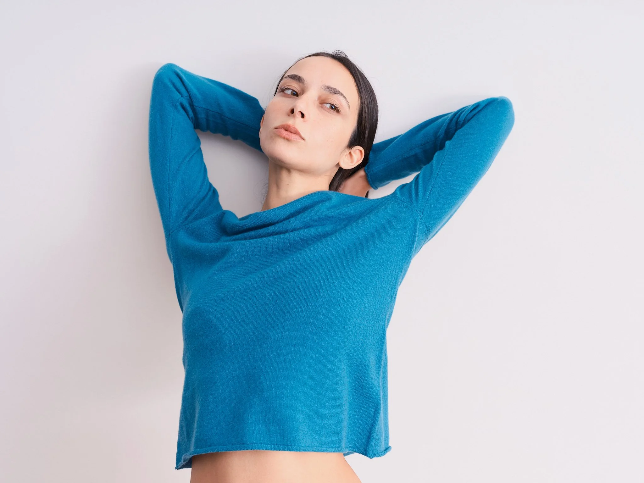 Mulher com suéter de cashmere de gola redonda cor turquesa, com as mãos atrás da cabeça, olhando para o lado, contra fundo branco.