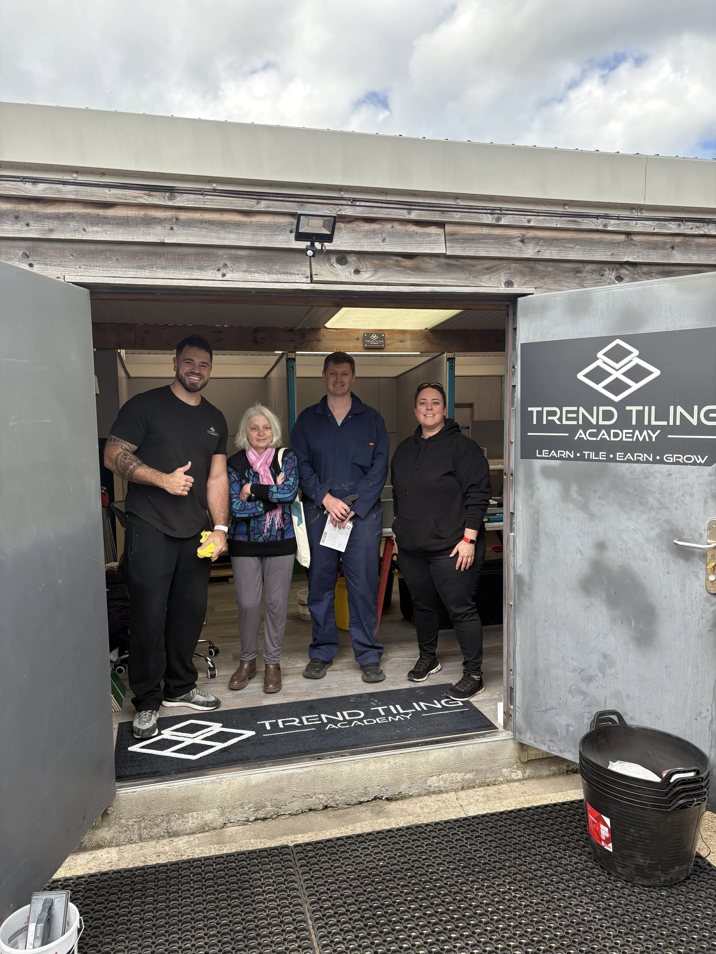Four people standing inside a door frame at Trend Tiling Academy, smiling at the camera. There are two men and two women, with one woman elderly. The background shows a workshop with tools and materials.