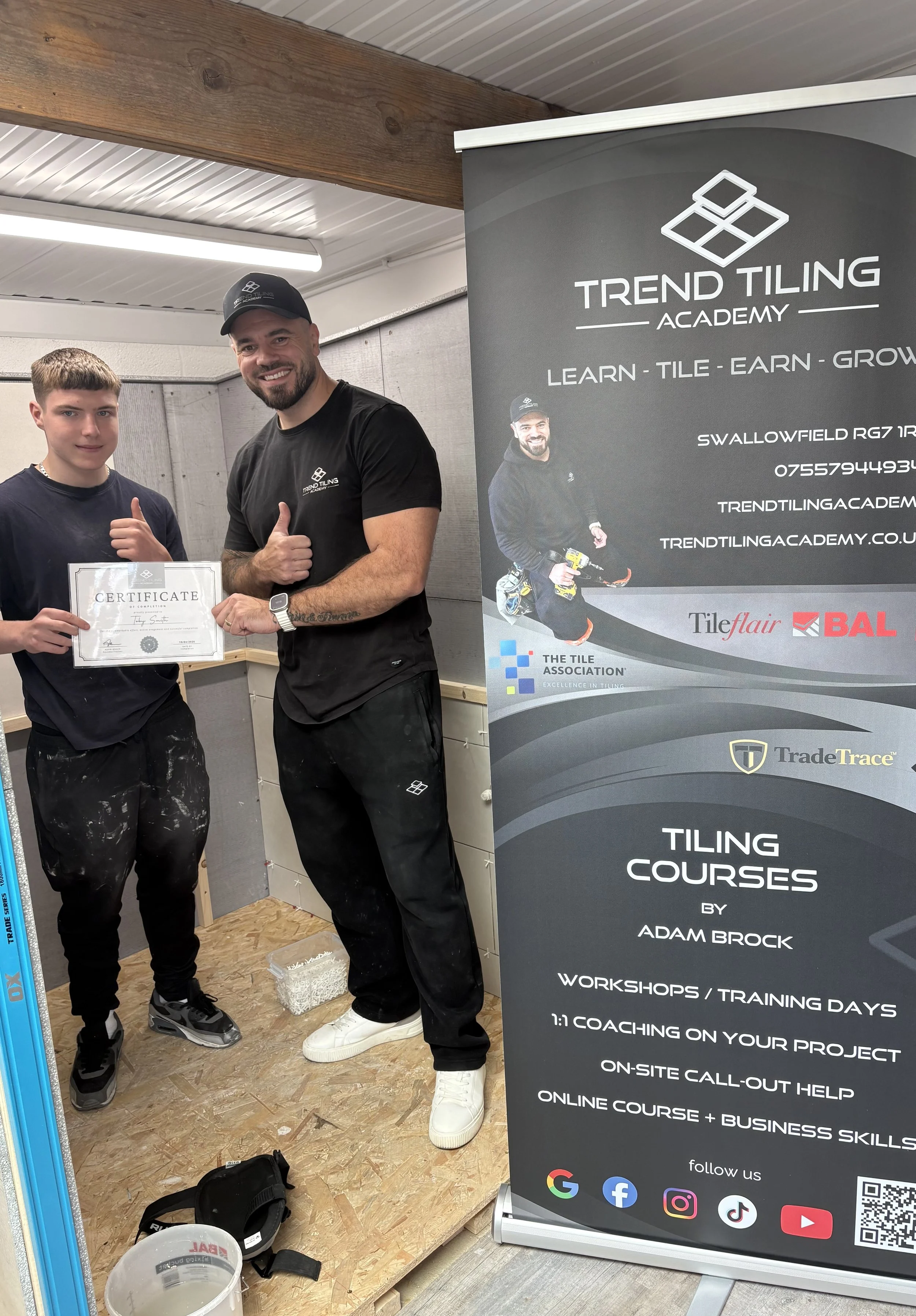 A man and a young boy standing inside a workshop, smiling and giving thumbs-up while holding a certificate. Next to them is a gray banner promoting tiling courses.