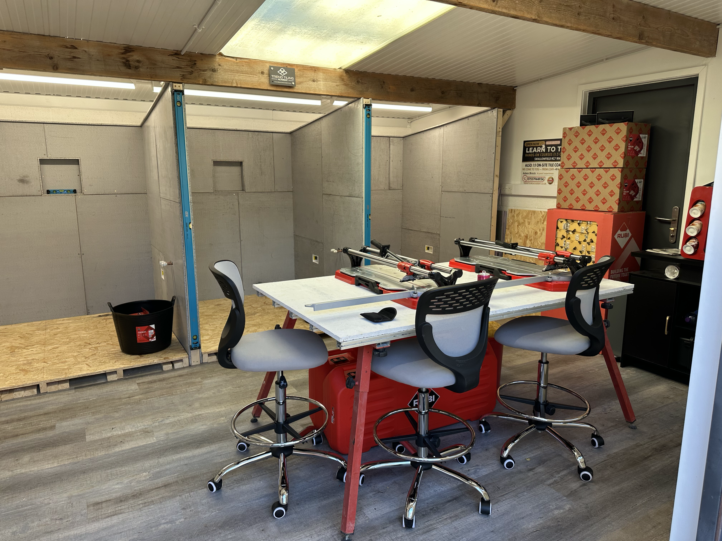 An indoor space under construction or renovation with unfinished walls and insulation panels, a white sheet of material on the ceiling, exposed wooden beams, and a white table with three black chairs. There are boxes of supplies, a fire extinguisher,