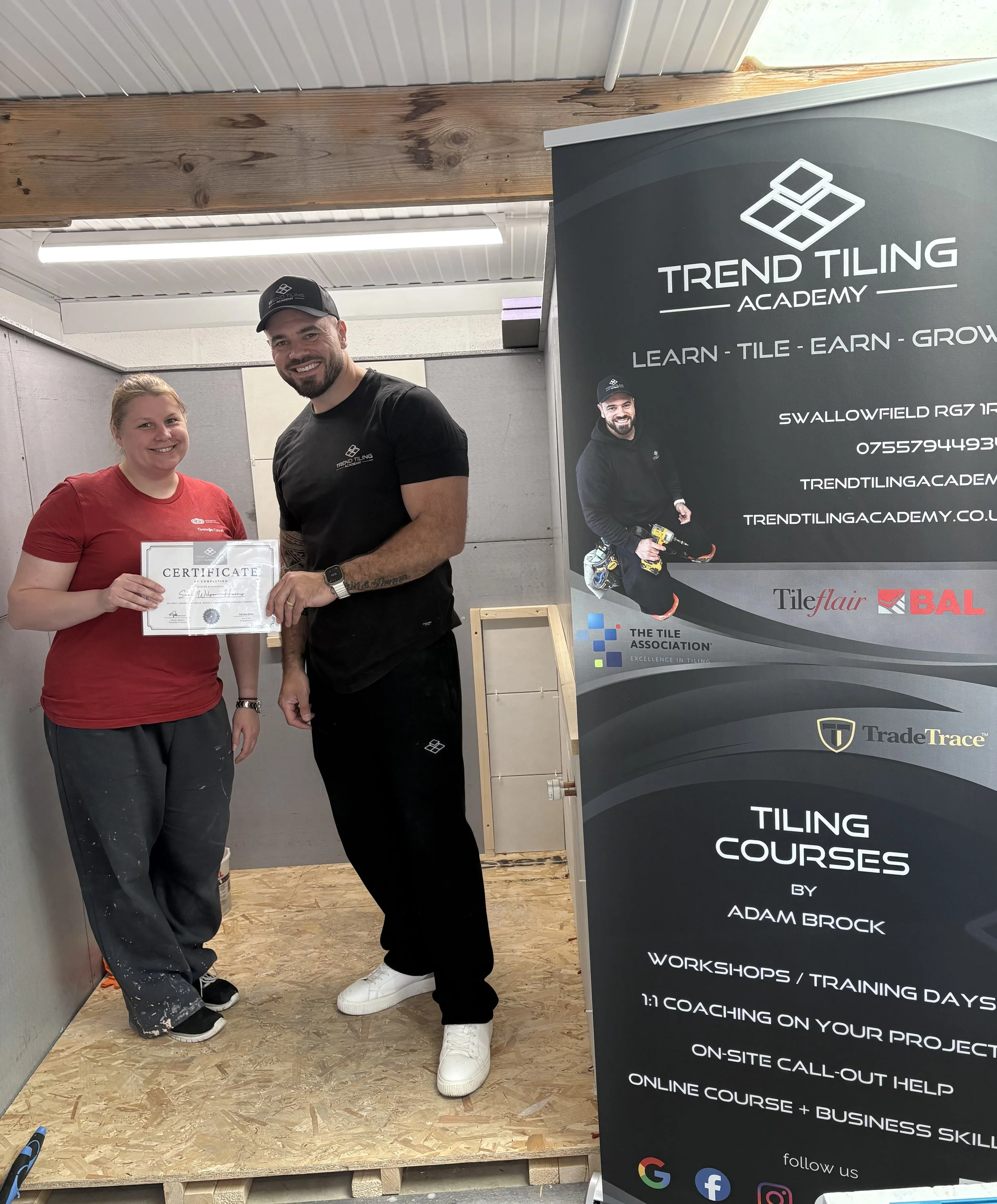 Two smiling people, a woman in a red shirt and a man in a black shirt and cap, standing indoors next to a large banner promoting tiling courses by Adam Brock at Trend Tiling Academy. The woman holds a certificate, and both are posing for the photo.