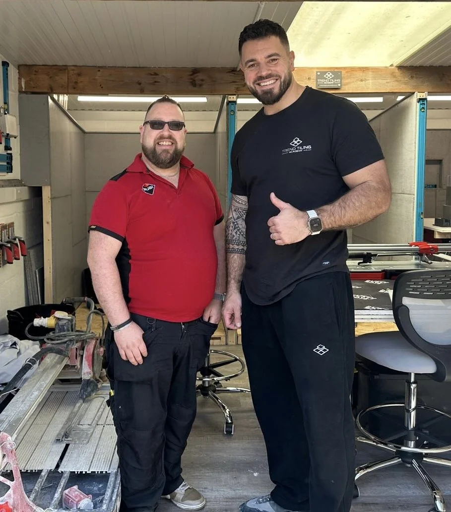 Two men smiling and standing together in a workshop or office, one wearing a red shirt and sunglasses, the other wearing a black shirt and giving a thumbs-up.