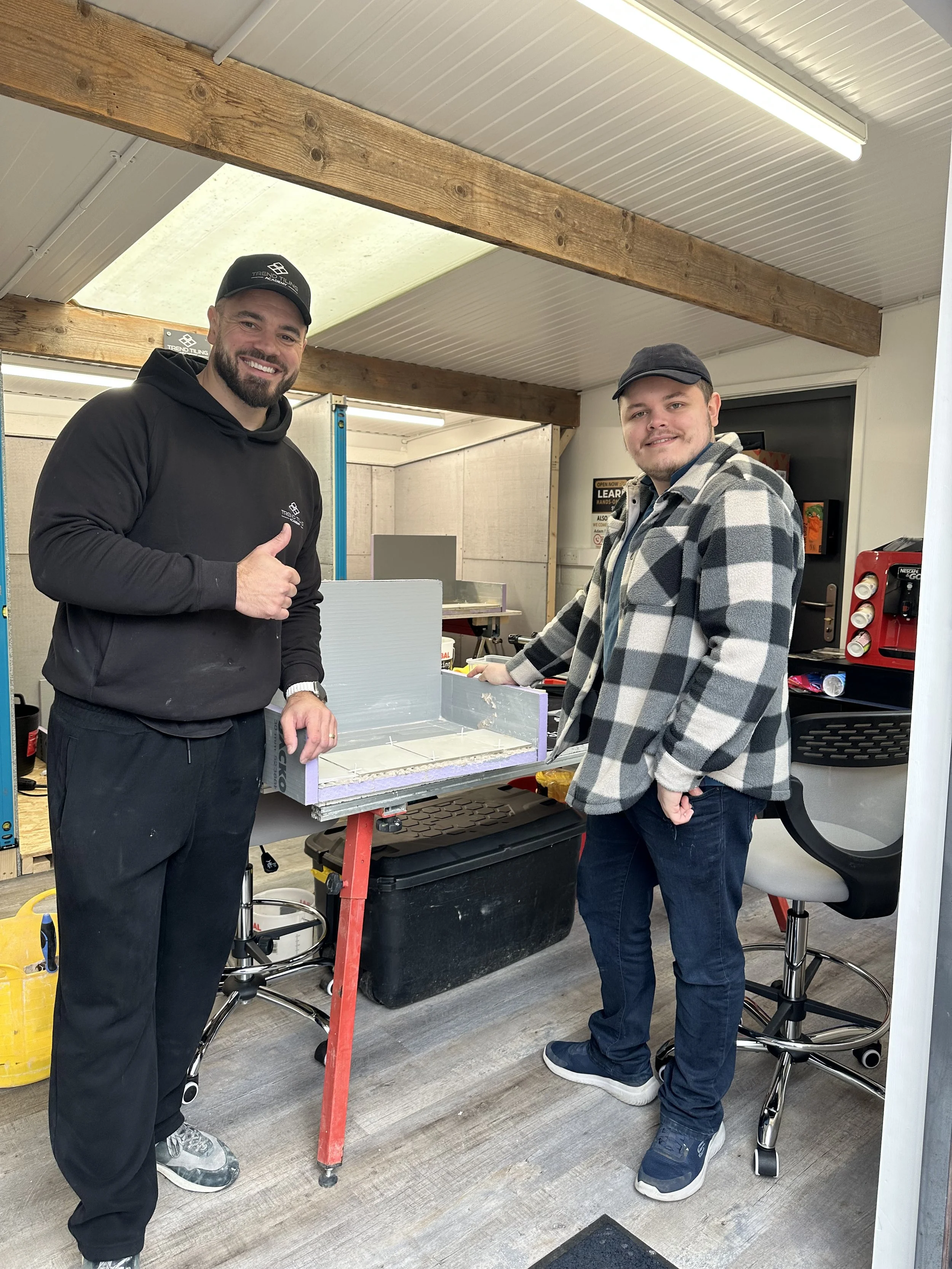 Two men are standing next to each other in a room with a workbench. One man, on the left, is smiling and giving a thumbs-up. He is wearing a black hoodie, black pants, sneakers, and a black cap. The other man, on the right, is also smiling, wearing a