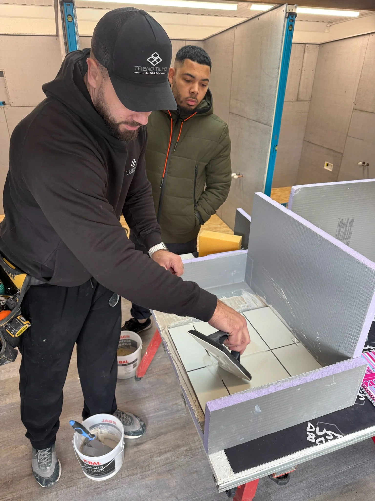 Two men working on a tiling project inside a room, with one applying adhesive on the floor and the other observing. The room's walls are partially tiled, and construction tools and materials are visible.
