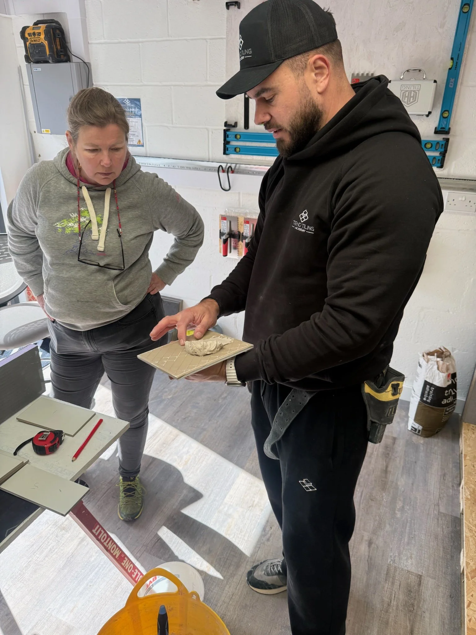 Two people working on a project in a workshop, inspecting a material or component on a small beige tray, with various tools and supplies around them.