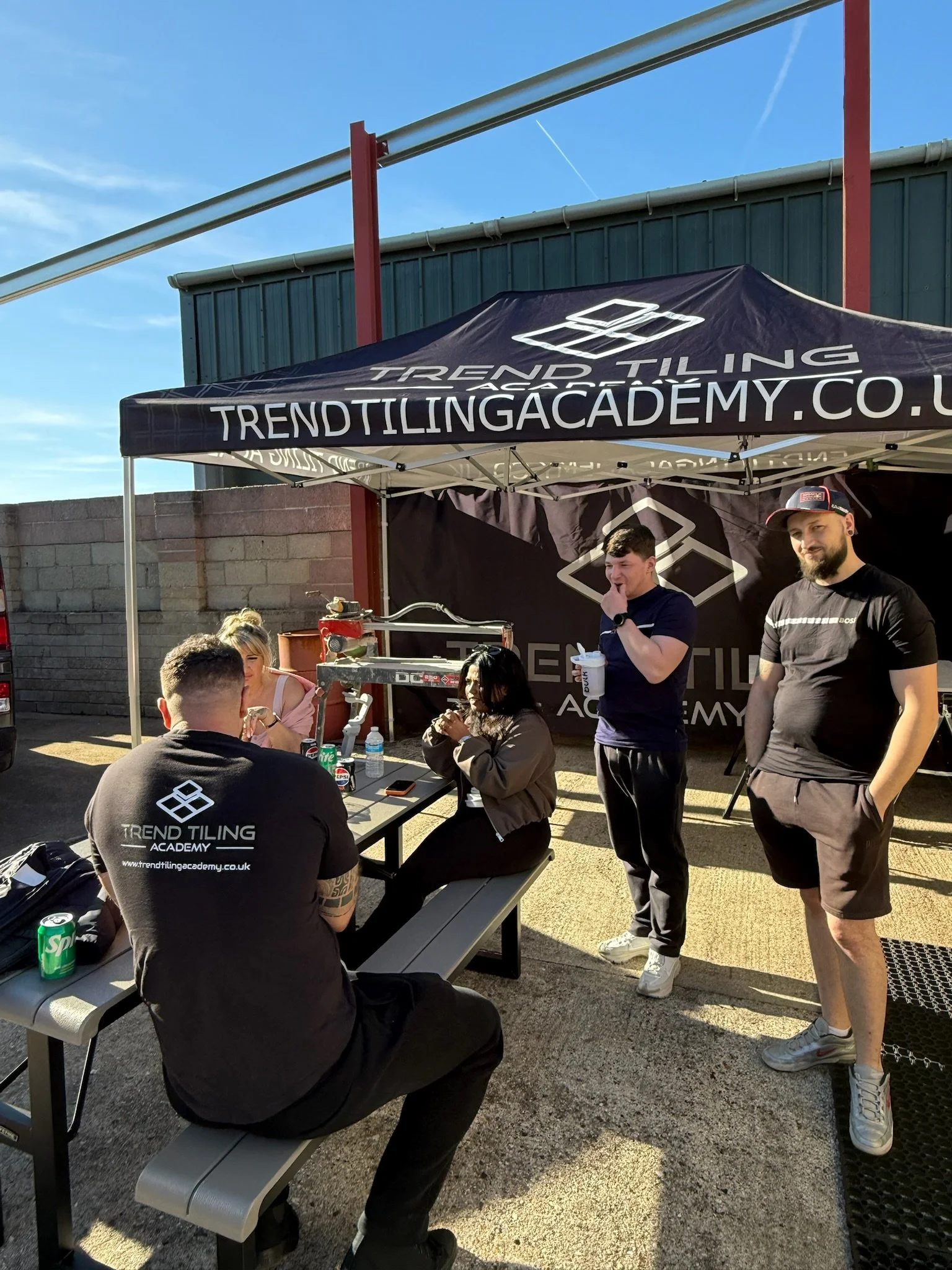 A group of people gathered under a black canopy tent with branding for Trend Tiling Academy. One person is seated at a table with beverages, while two others stand nearby, engaged in conversation. The setting appears to be outdoors, with a brick wall