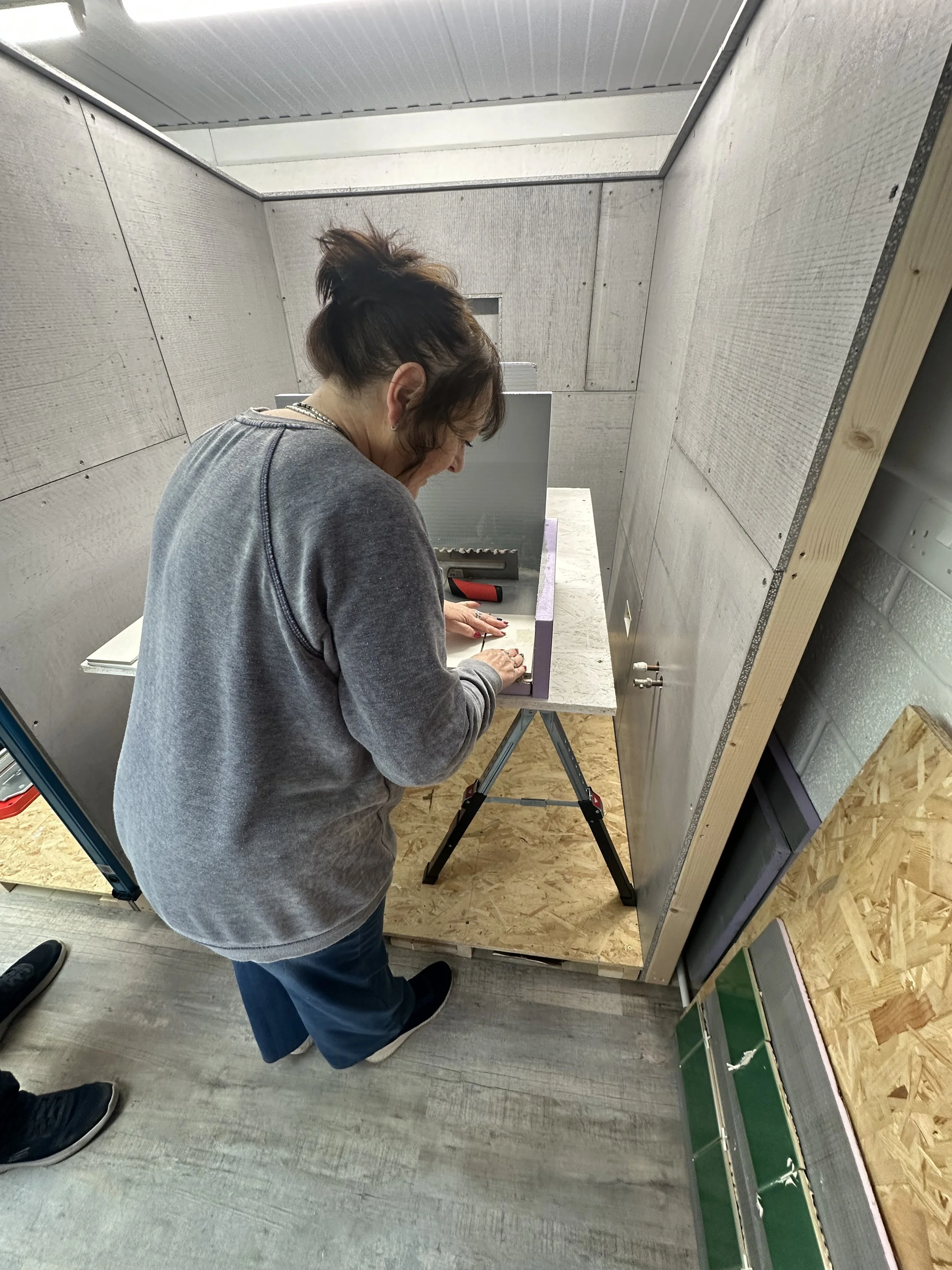 A woman with dark hair in a messy bun, wearing a gray sweatshirt and necklace, is working on a small project at a table with a purple border and a gray surface in a woodworking or crafting space. The space has gray and wood-paneled walls and ply floo