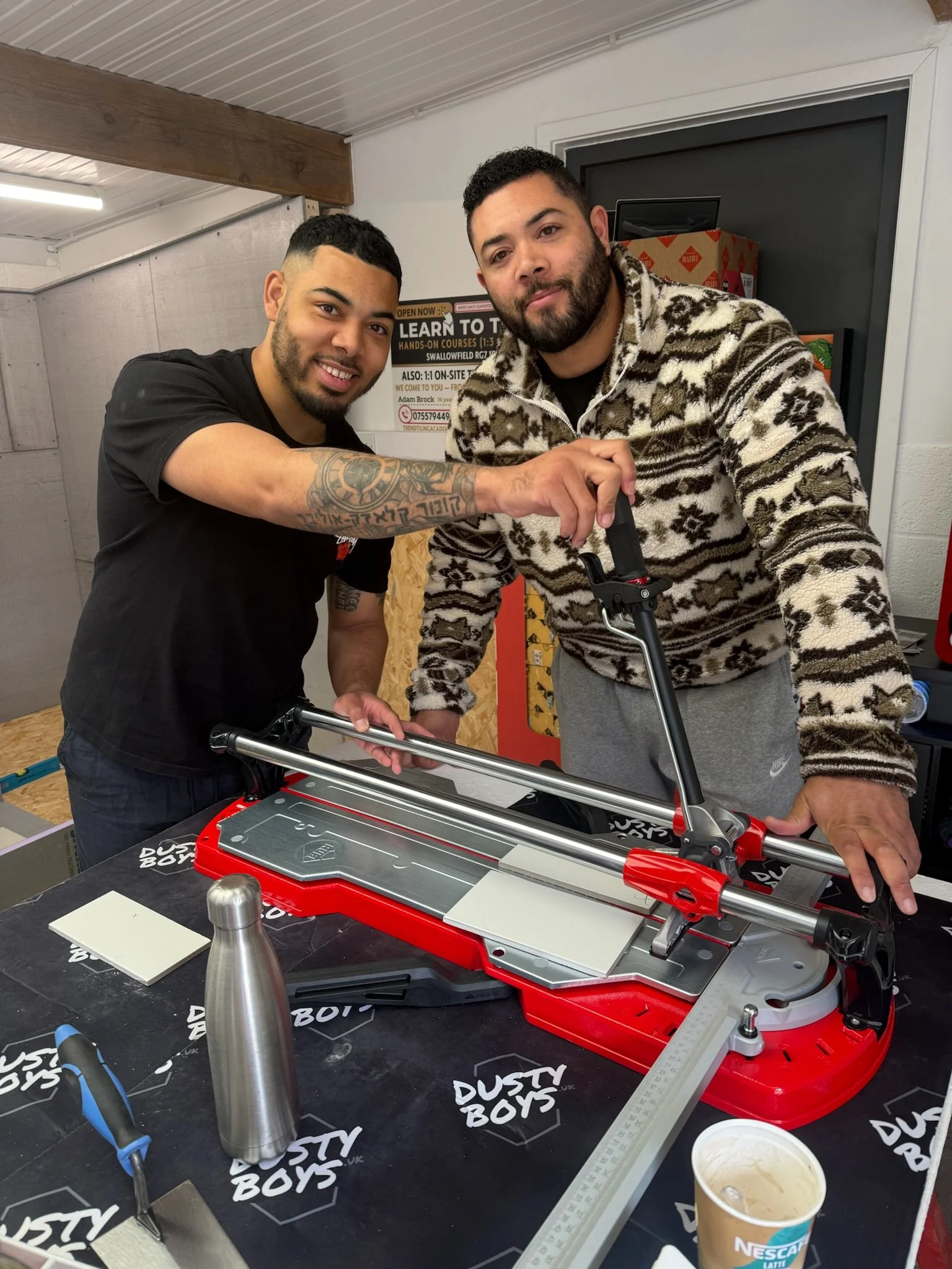 Two men are working together on a mechanical project, with one holding the other’s arm, in a workspace with tools, a ruler, and a tub of ice cream on the table.