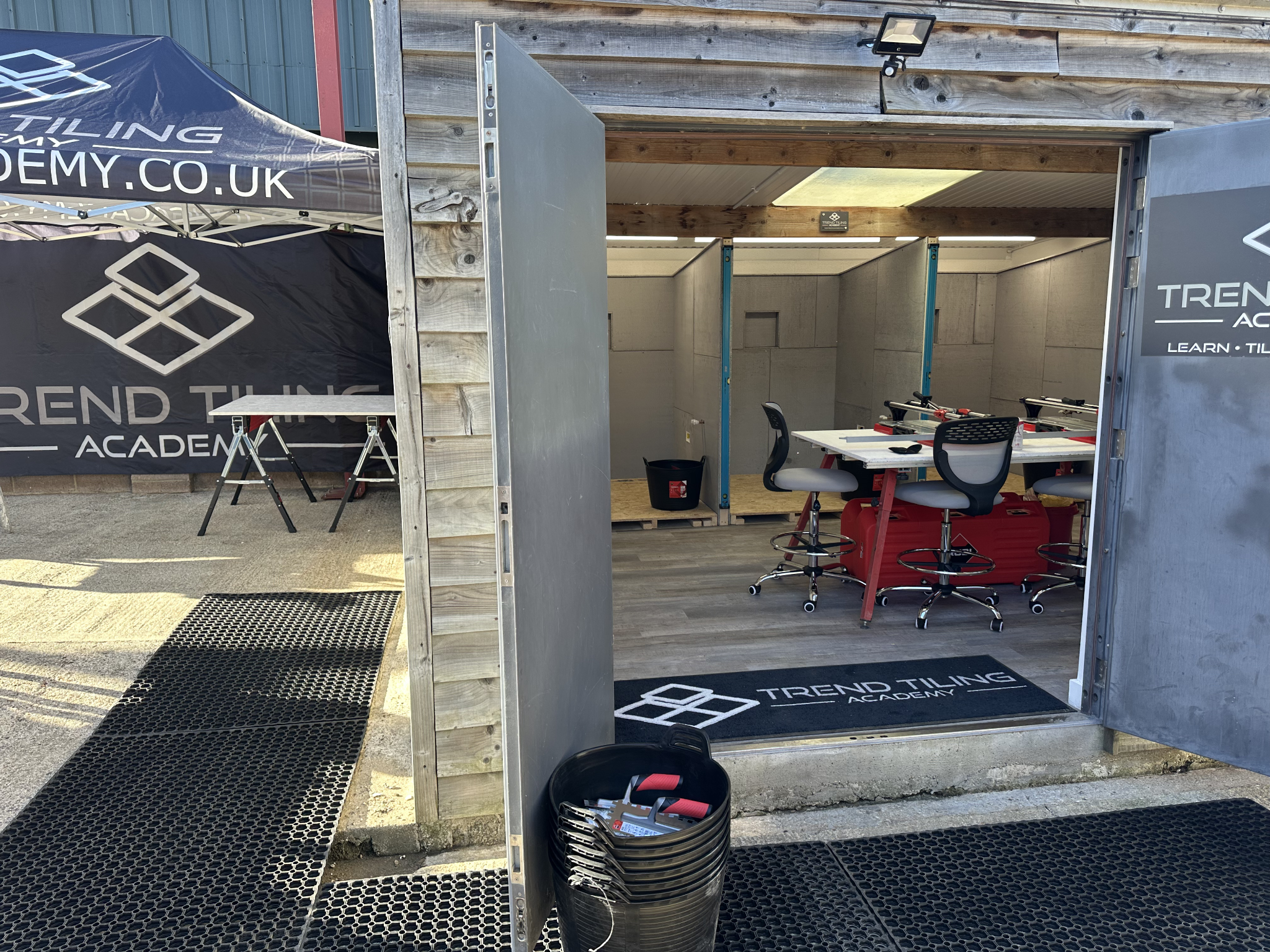 A partially constructed indoor space with wooden and metal framing, desks, office chairs, and black trash bins, with branding for "Trend Tiling Academy" on a nearby tent and a mat.