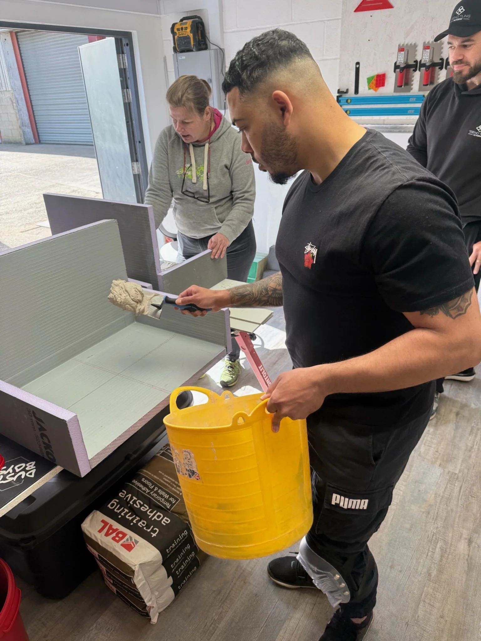 Three people working together in a workshop. One person is holding a yellow bucket and a cutter, working on a project that involves insulation material. The other two individuals are observing and assisting near a work table with supplies and tools. 