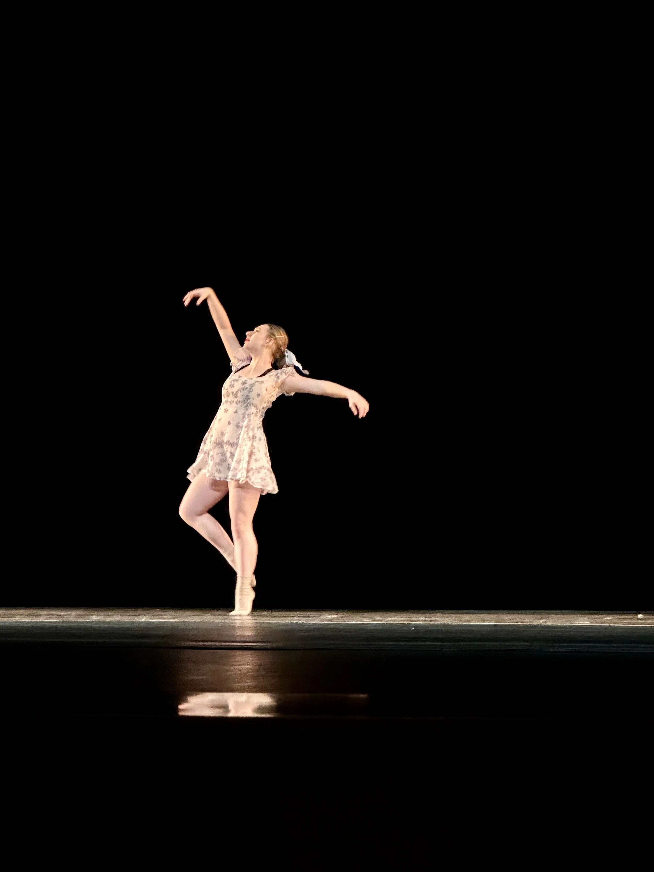 Jacksonville teen ballet dancer performing on stage with her arms raised gracefully, wearing a floral dress, against a black background.