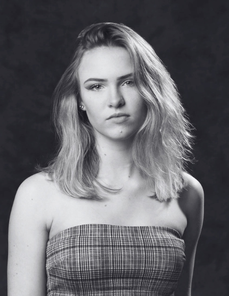 Black and white portrait of a young woman with shoulder-length wavy hair, wearing a strapless checkered top, looking at the camera with a serious expression.