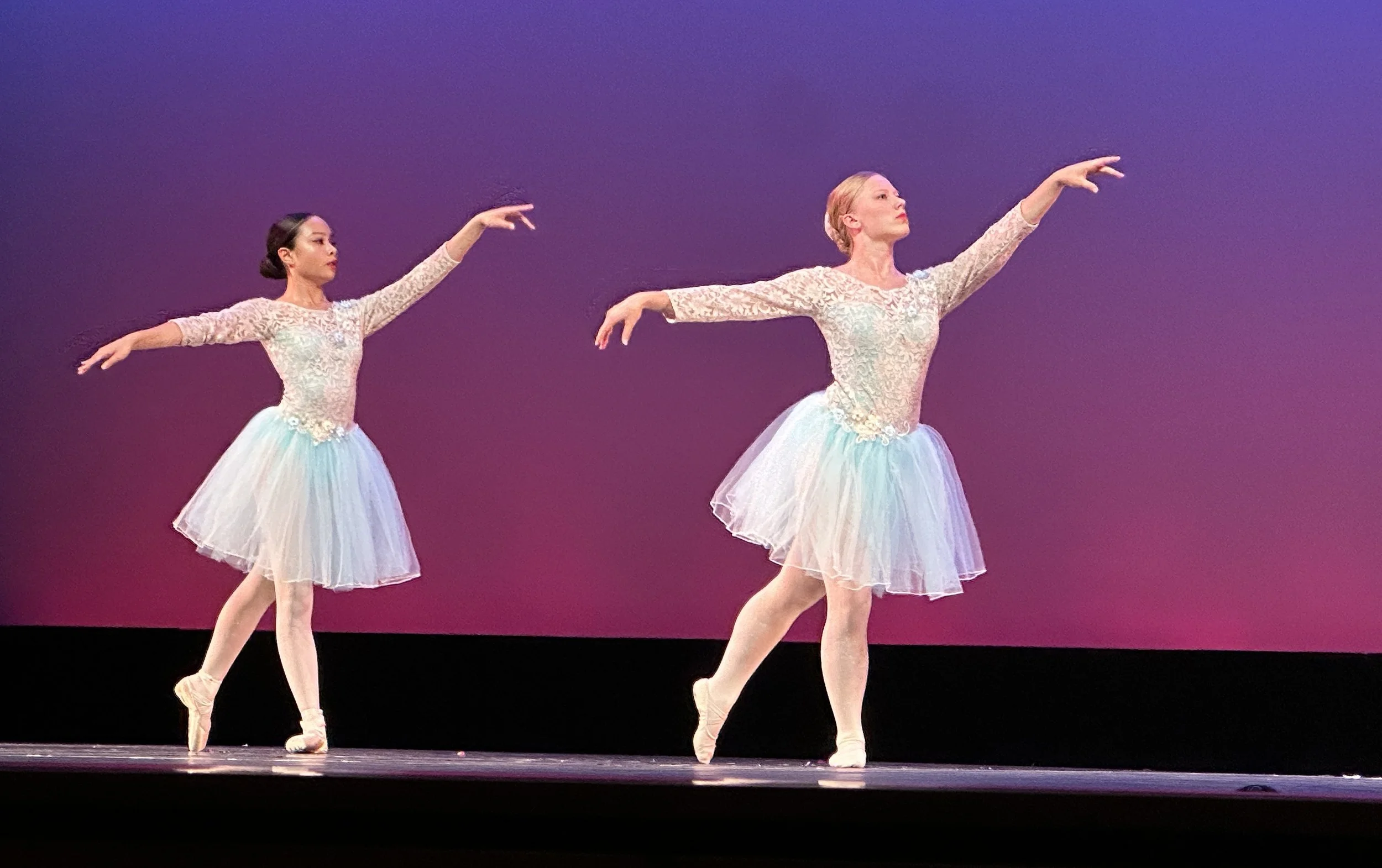 Two advanced ballet dancers in lace costumes performing on stage at recital with a purple background.
