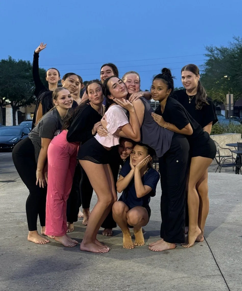Group of nine young women and girl Jacksonville dance studio students posing outdoors during evening, some hugging and smiling, with a clear sky and parked cars in the background.