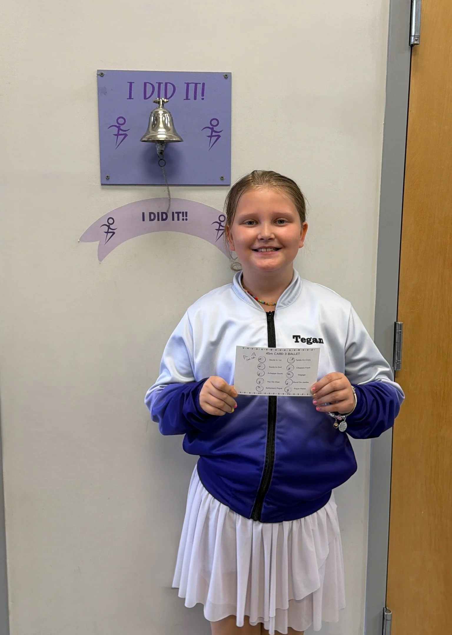 A young girl with brown hair in a ponytail, smiling and holding a piece of paper, standing in front of a wall with a purple sign that says 'I DID IT!' and a small bell above it. She is wearing a white jacket a pleated white skirt.