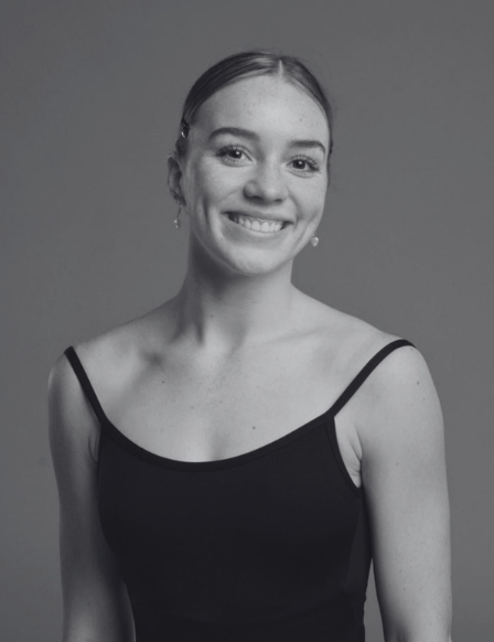Black and white portrait of a young woman smiling, wearing a sleeveless top with thin straps and earrings, against a plain gray background.
