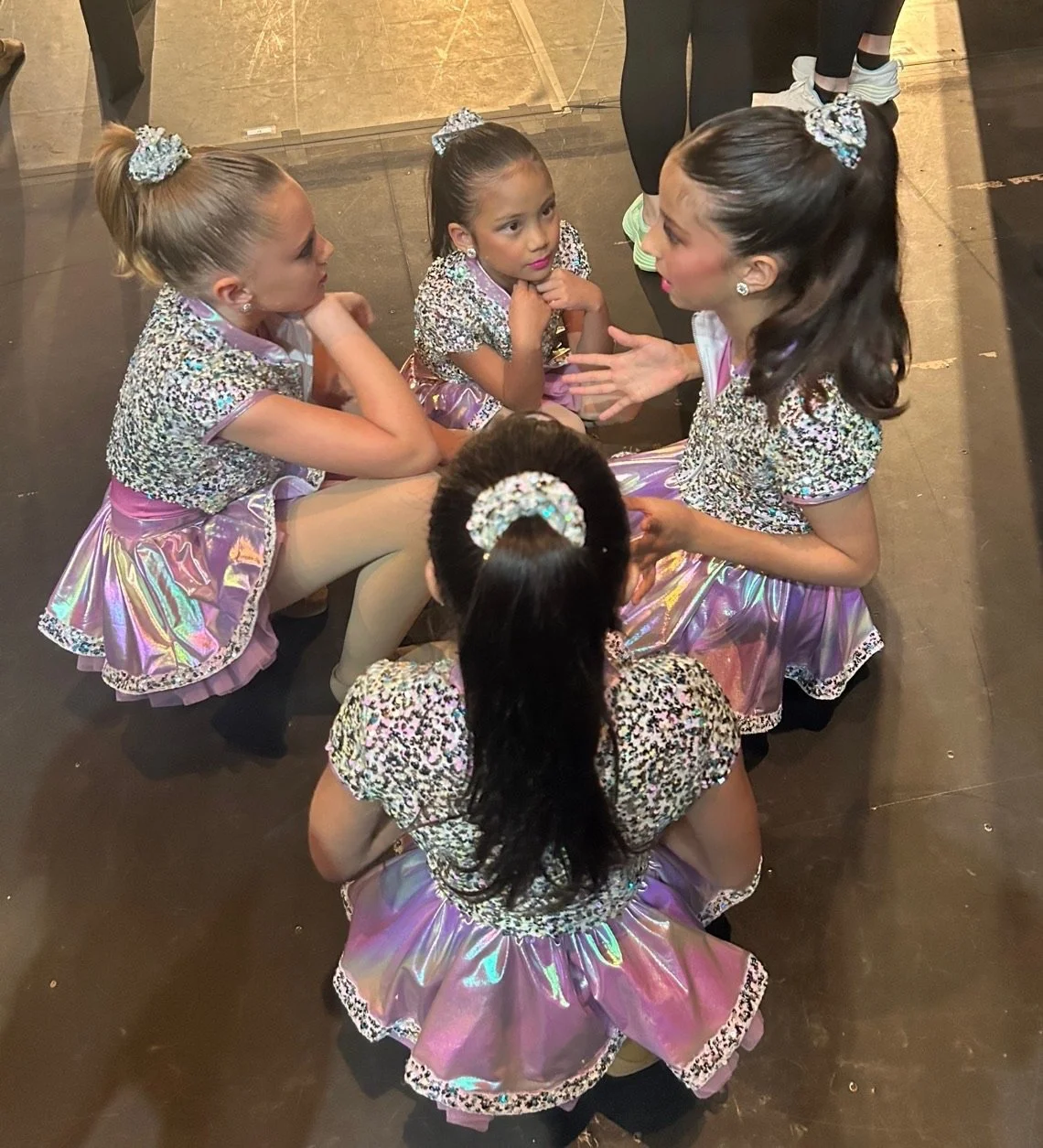 Five young girls dressed in matching shiny, glittering costumes with leopard print and pink accents, sitting in a circle and engaging in a conversation backstage at a dance competition.