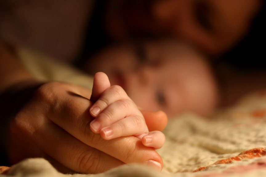 A close-up of an adult holding a baby's hand, with the baby lying in the background.