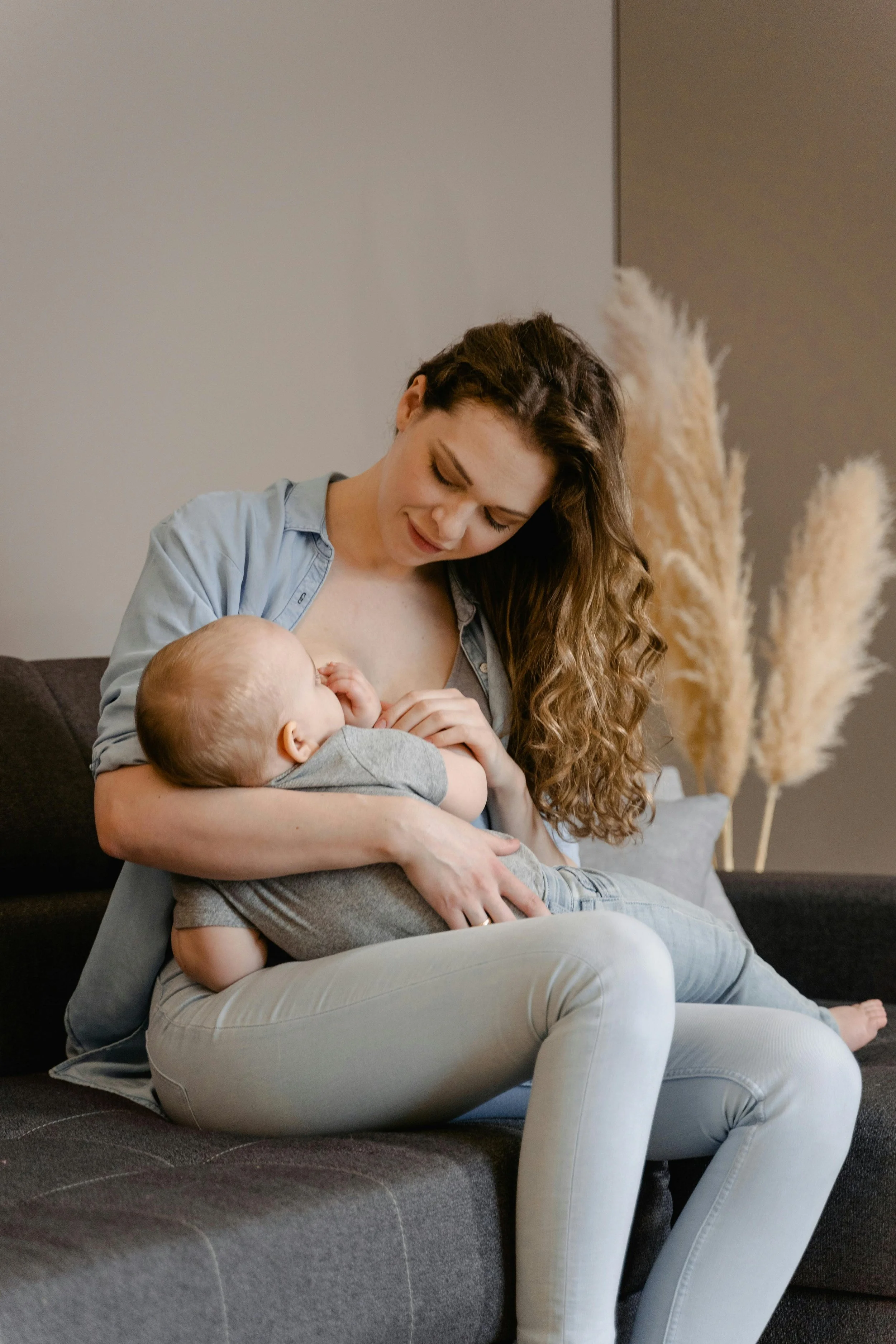 A woman nursing a baby on her lap while sitting on a couch in a living room. The woman has long, wavy hair and is wearing a light blue shirt and light-colored pants. There is a large, fluffy pampas grass arrangement in the background.