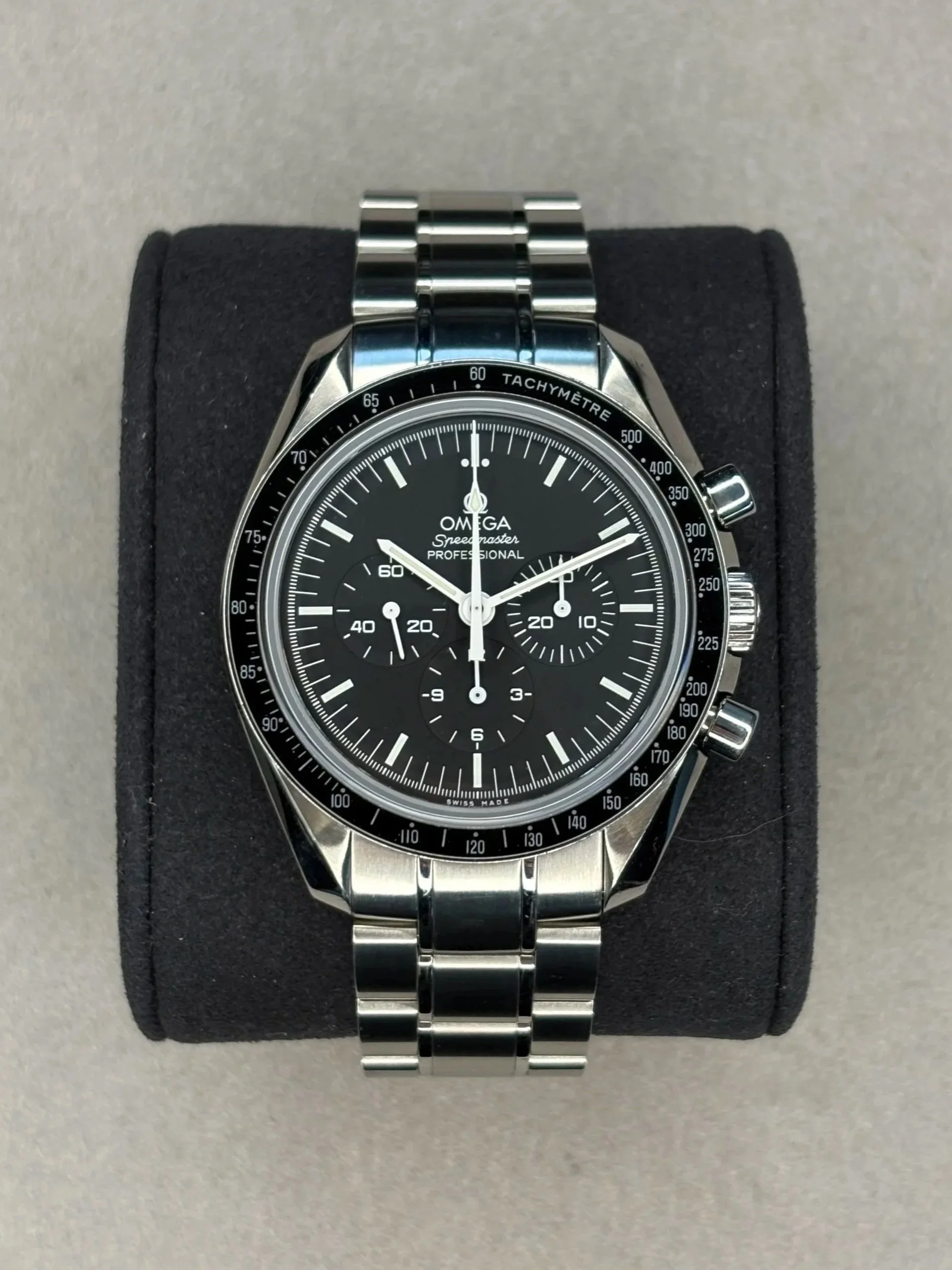 A stainless steel Omega Speedmaster Professional wristwatch with a black dial, three subdials, and a tachymeter bezel, displayed on a black cushion.