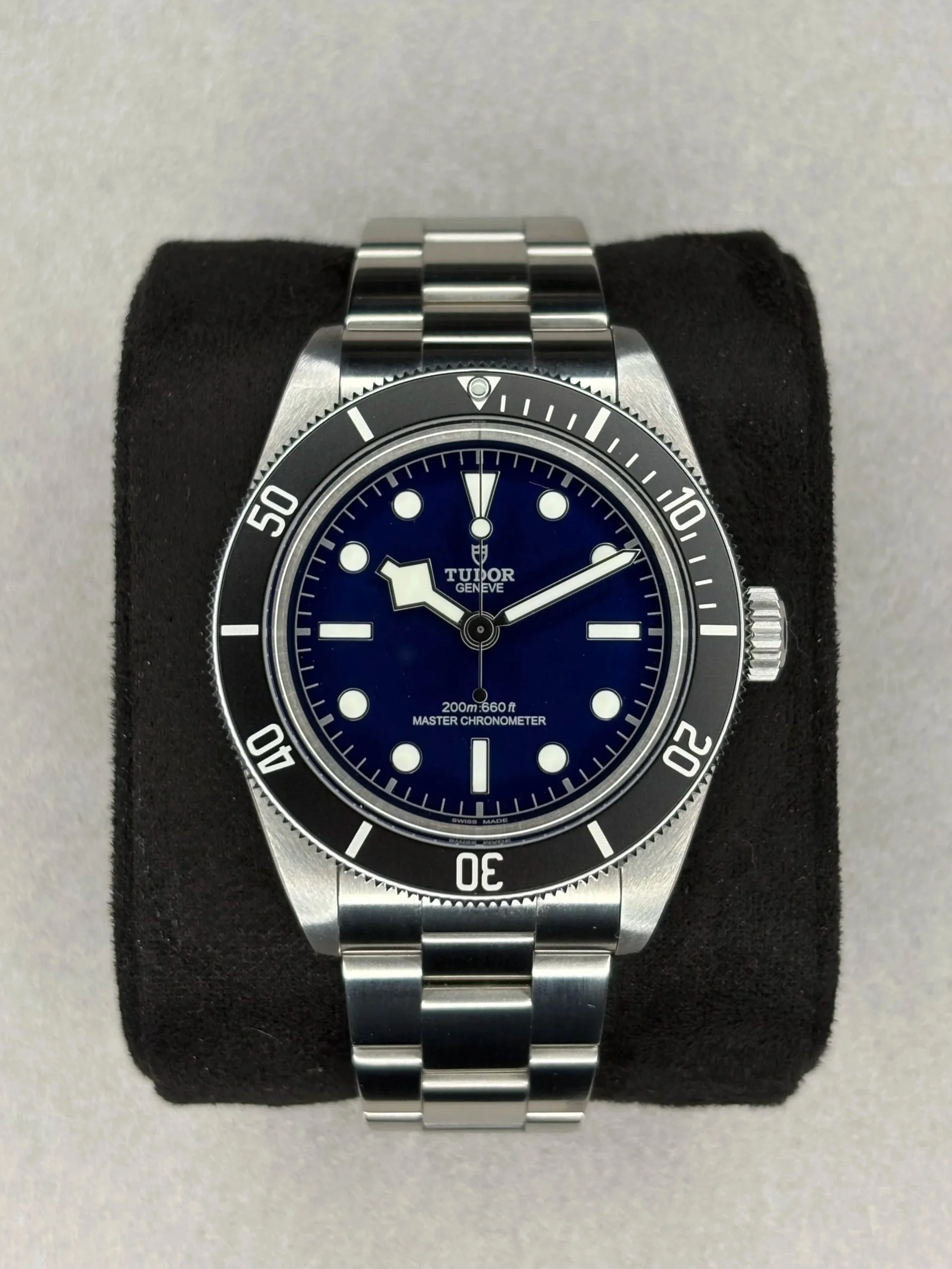 A Tudor dive watch with a blue dial, stainless steel bracelet, and black rotating bezel, displayed on a black cushion.