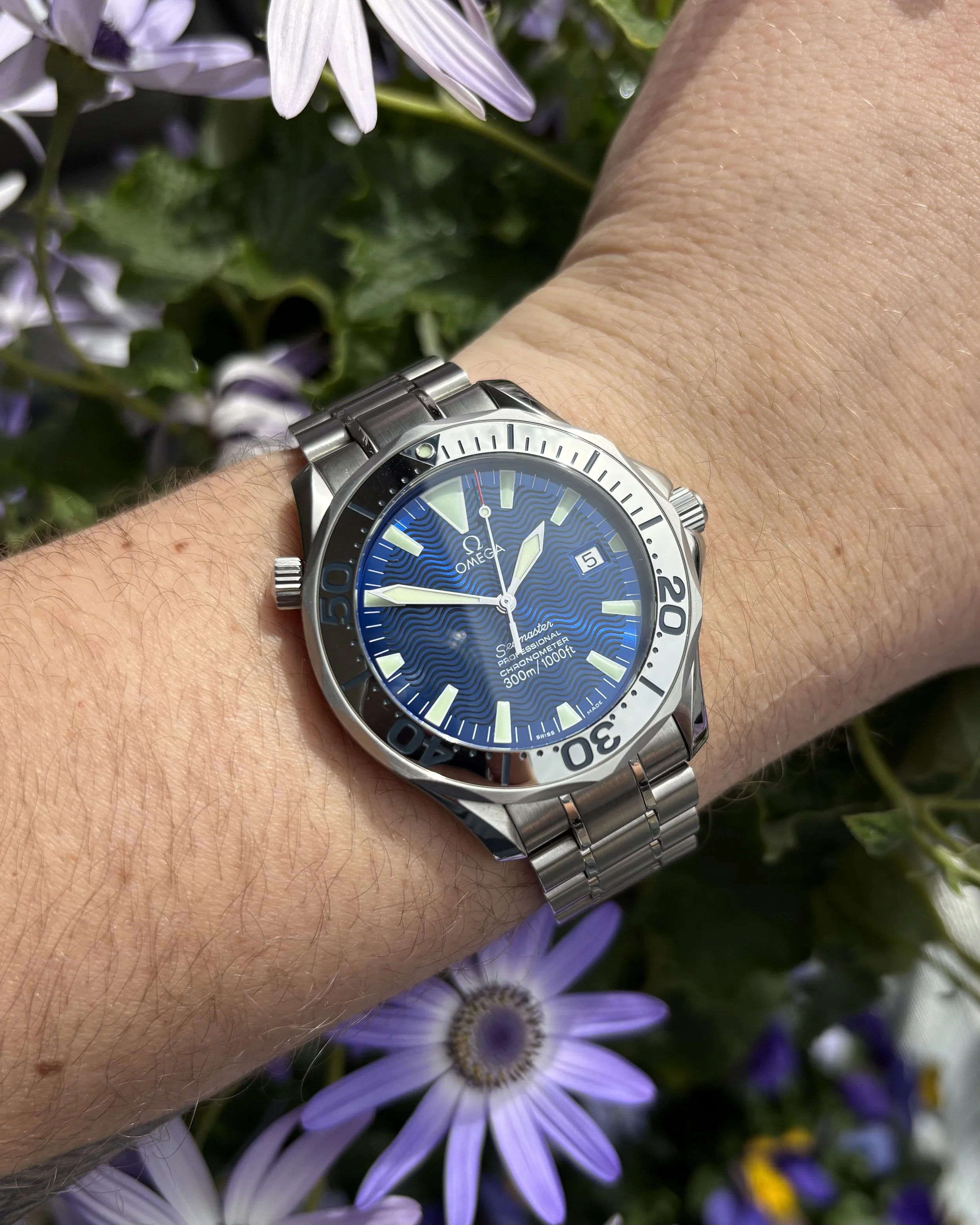 A wristwatch with a blue face and silver band on a person's wrist, surrounded by purple and white flowers.