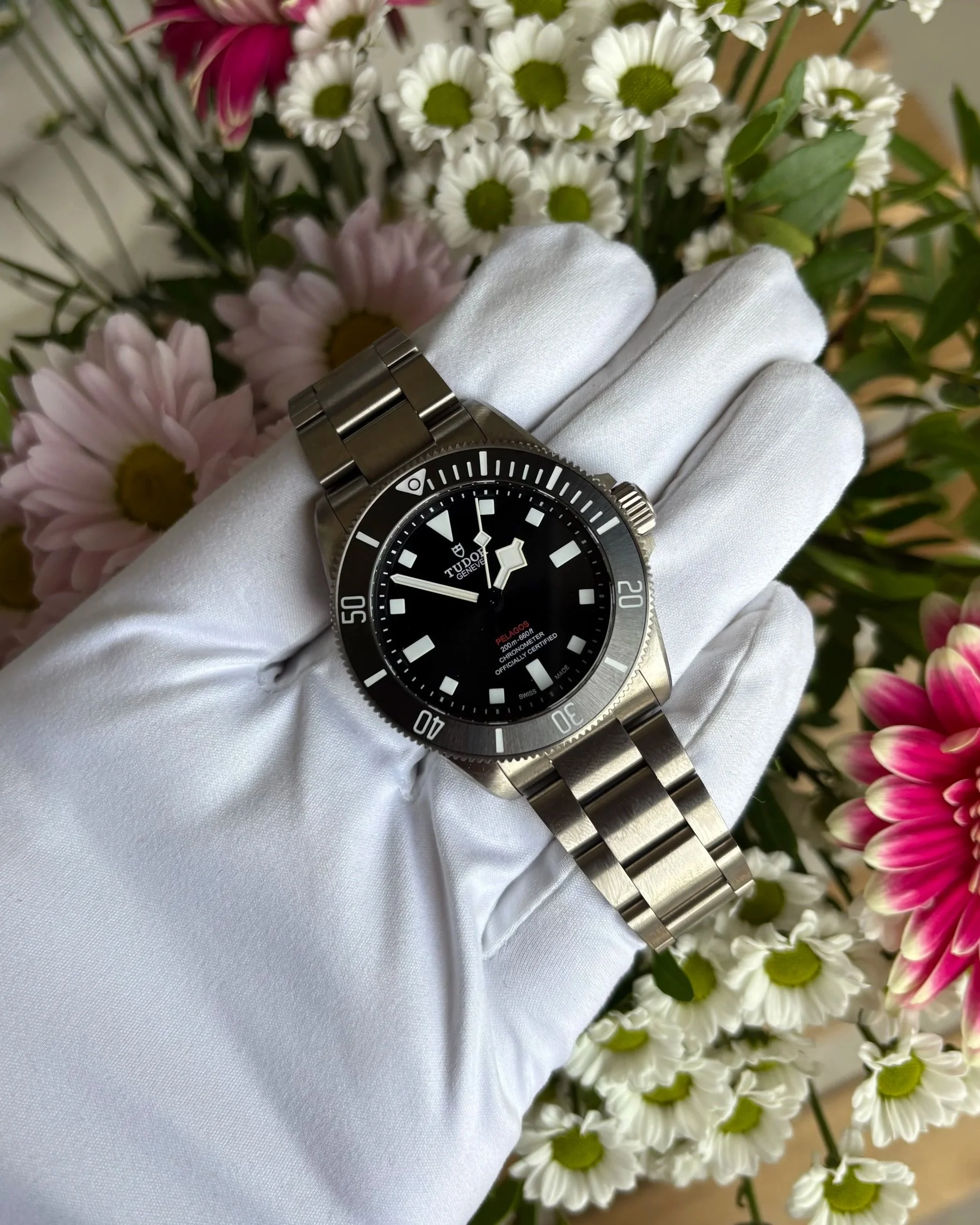 A person's hand dressed in a white glove displays a Tudor Pelagos diving watch in front of a bouquet of white daisies and pink flowers.