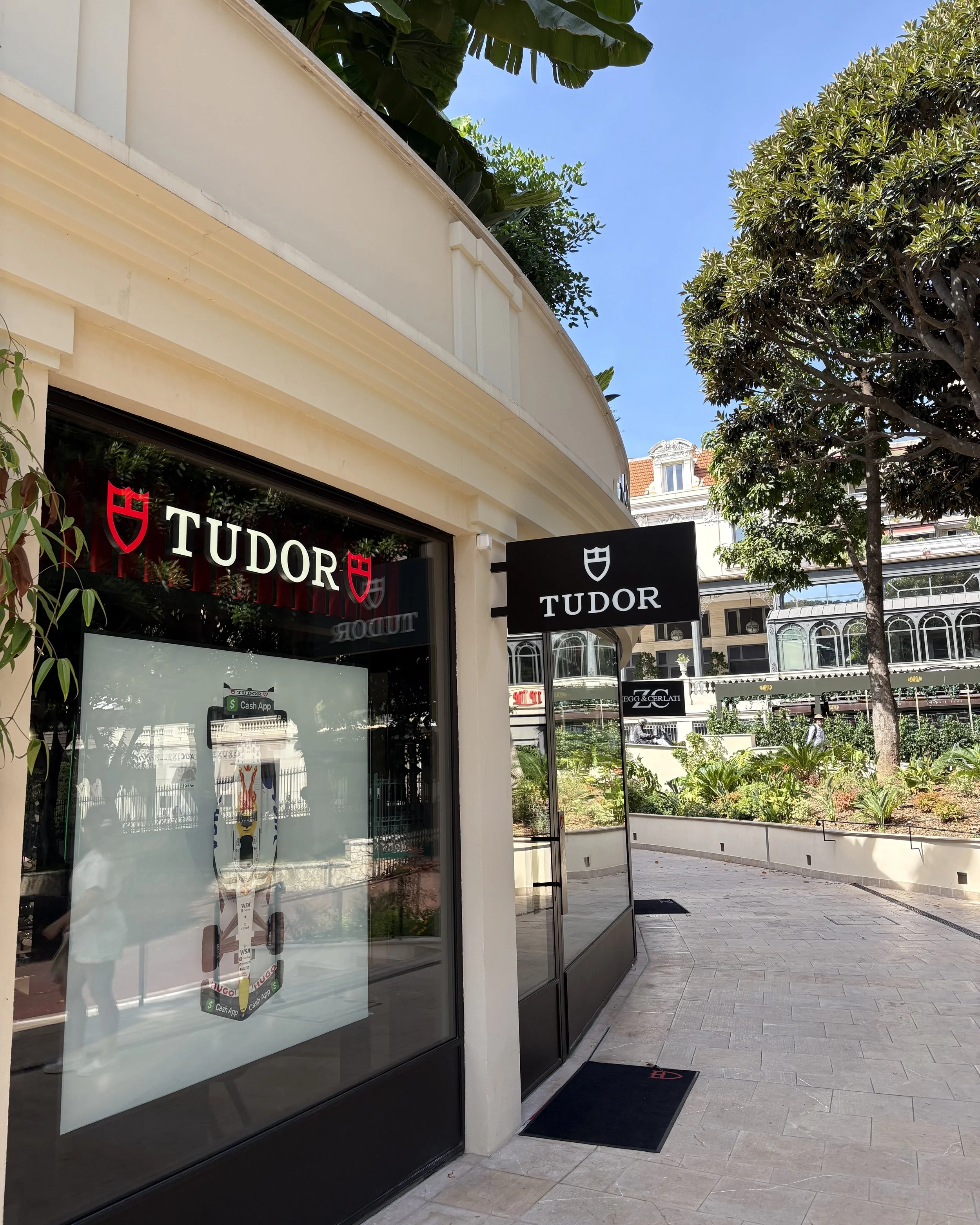 Exterior view of a Tudor jewelry store with large glass windows, black signage with a shield logo, and a sidewalk in front. Green trees and buildings are visible in the background on a sunny day.