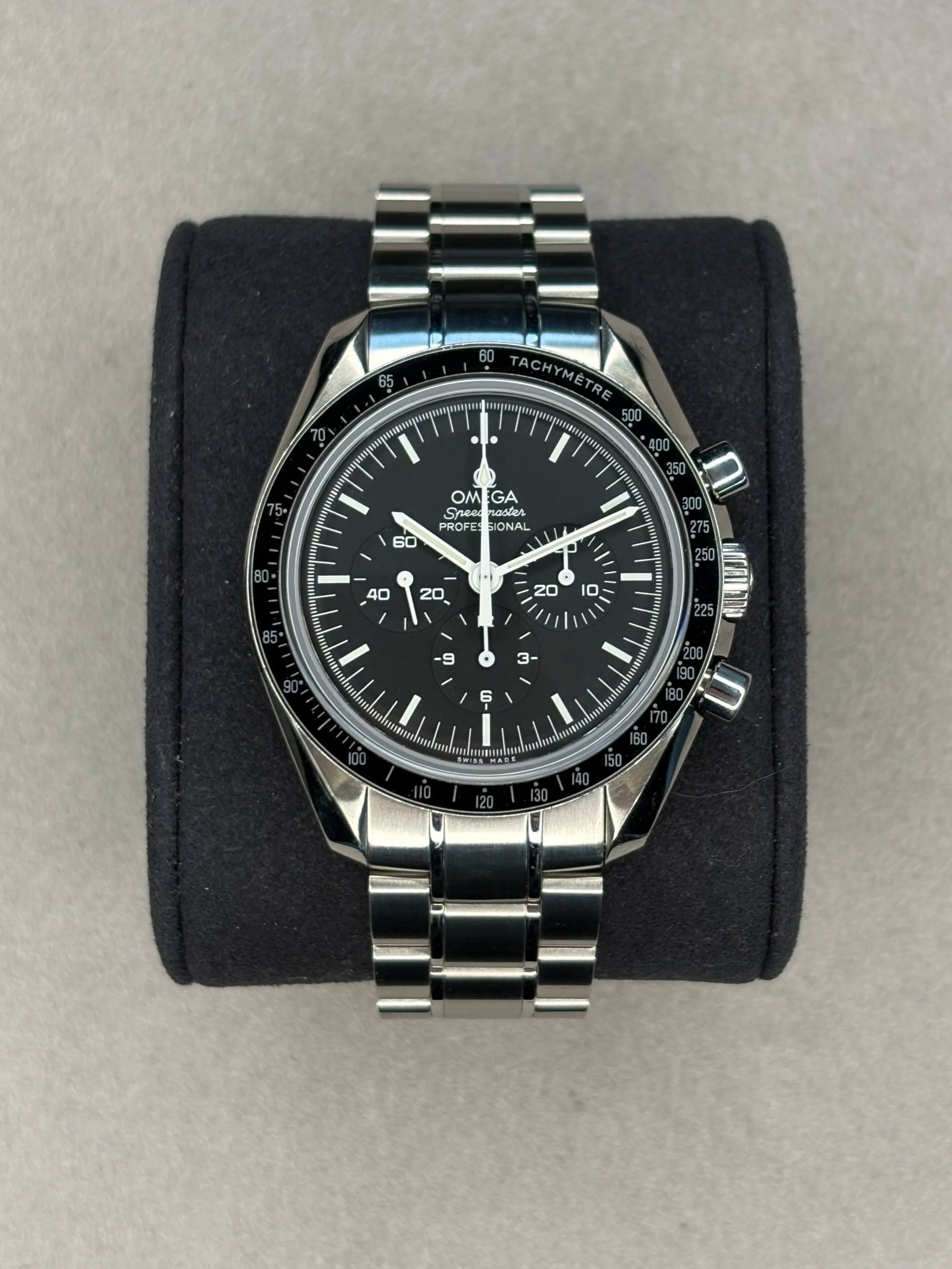 Close-up of an Omega Speedmaster Professional wristwatch with a metallic band, black dial, three sub-dials, tachymeter scale on the bezel, and chronograph pushers on the side, resting on a black cushioned display.