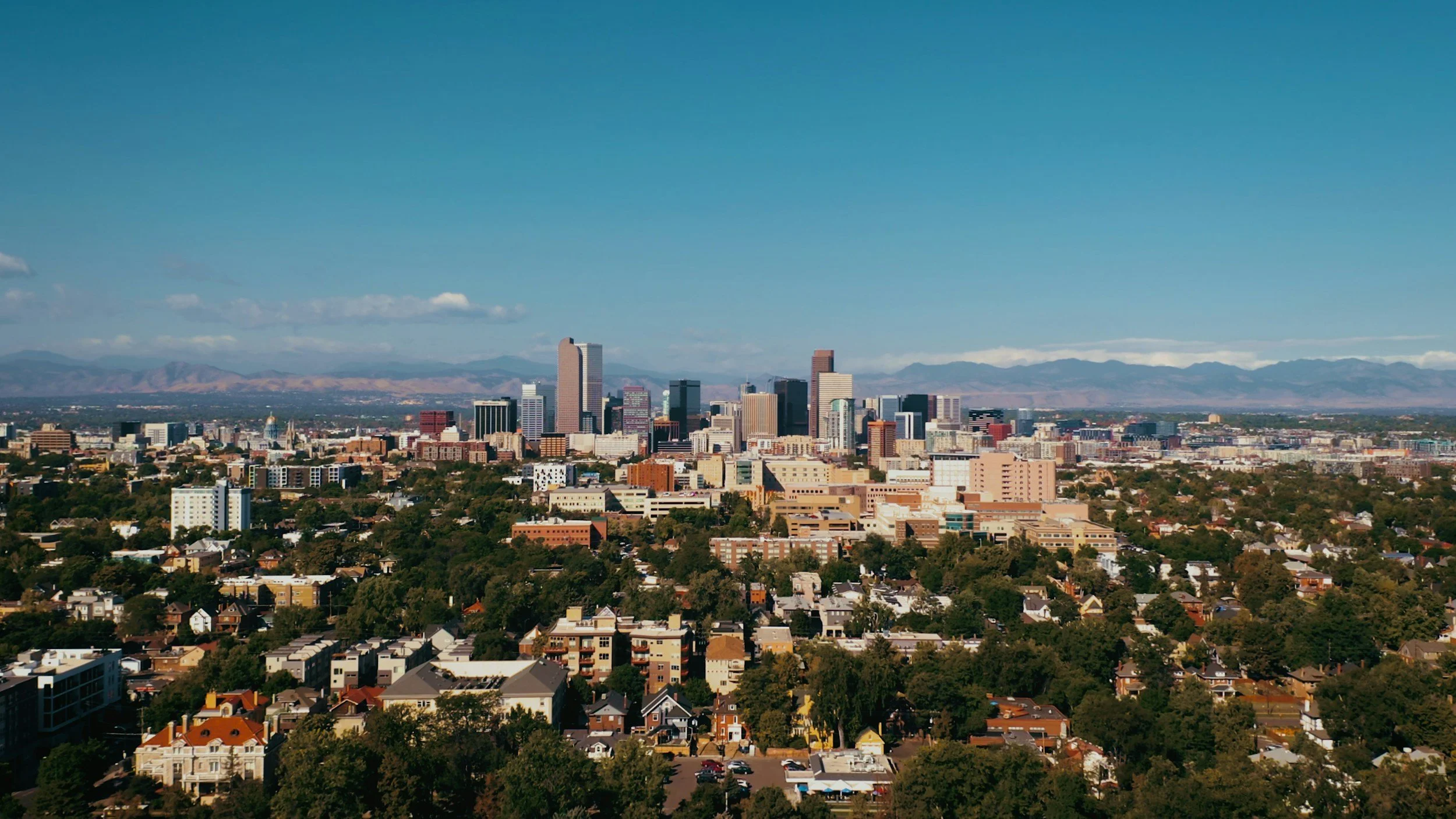 A city skyline with tall skyscrapers, residential neighborhoods in the foreground, and mountains in the background under a clear blue sky.