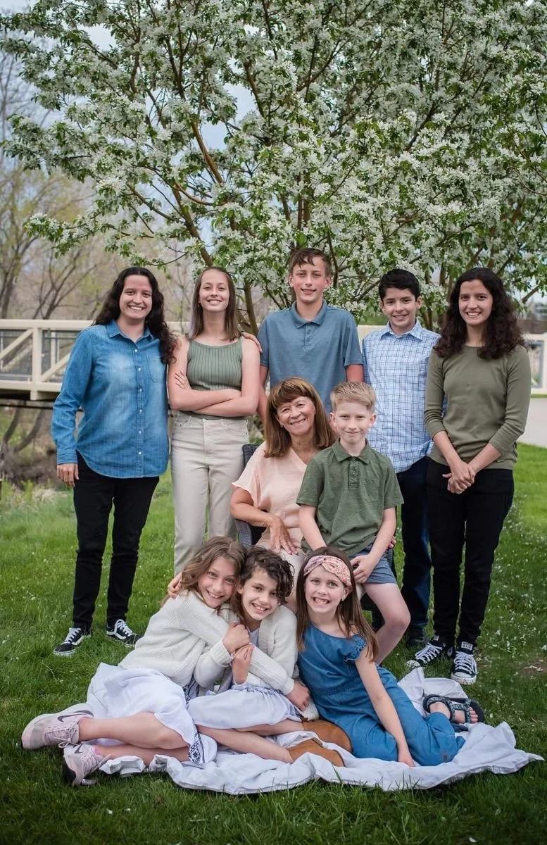A group of seven children and one adult woman sitting and standing outdoors in front of a blossoming tree. They are smiling and posing for a family photo on a grassy area.