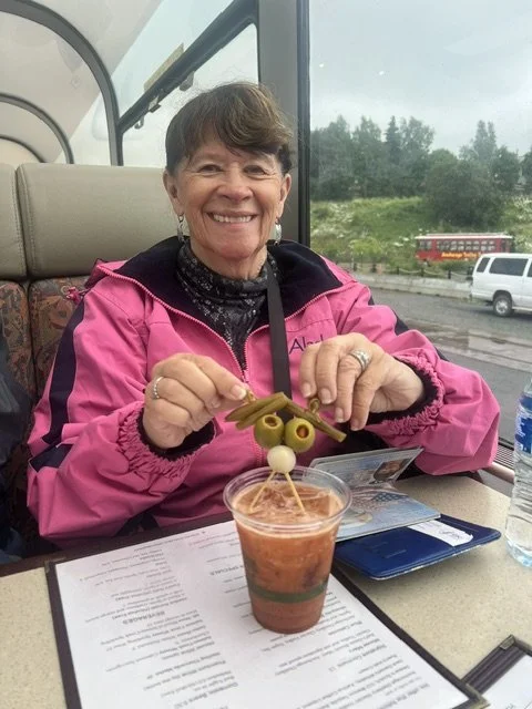 Smiling woman sitting on a bus, holding a skewer with olives and a pearl onion over a drink. There are travel documents on the table in front of her.