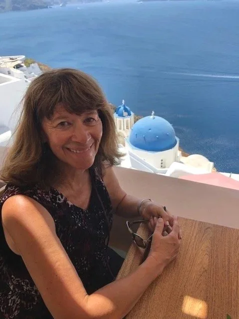 Smiling woman sitting at a dining table with a view of blue-domed buildings and the ocean in Santorini, Greece.