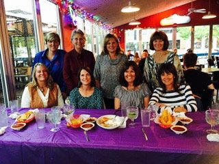 Group of eight women gathered around a table with food at a restaurant decorated with balloons and streamers.