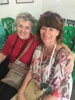 Two smiling women sitting together on green plastic chairs at what appears to be an indoor event, with one woman dressed in a red top and the other in a patterned blouse and glasses.