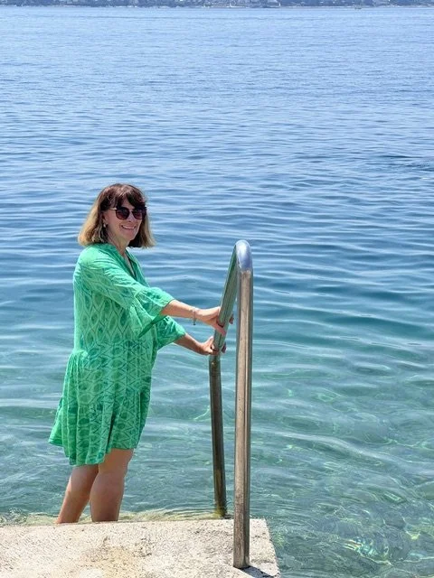 Woman in a green dress with sunglasses standing in clear blue water holding a metal handrail at a beach or lakeside.