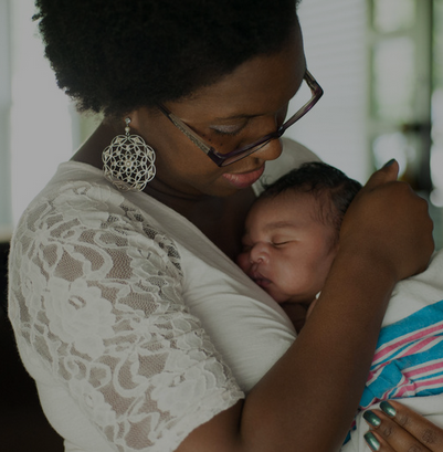 Black woman with glasses and earrings holding a sleeping baby wrapped in a blanket.