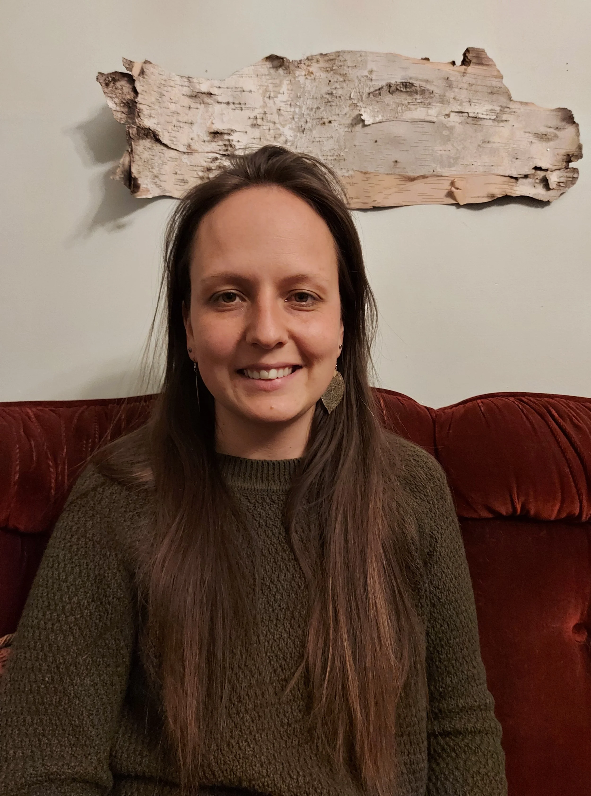A woman with long brown hair smiling, sitting on a red velvet sofa, with a piece of birch bark mounted on a plain wall behind her.