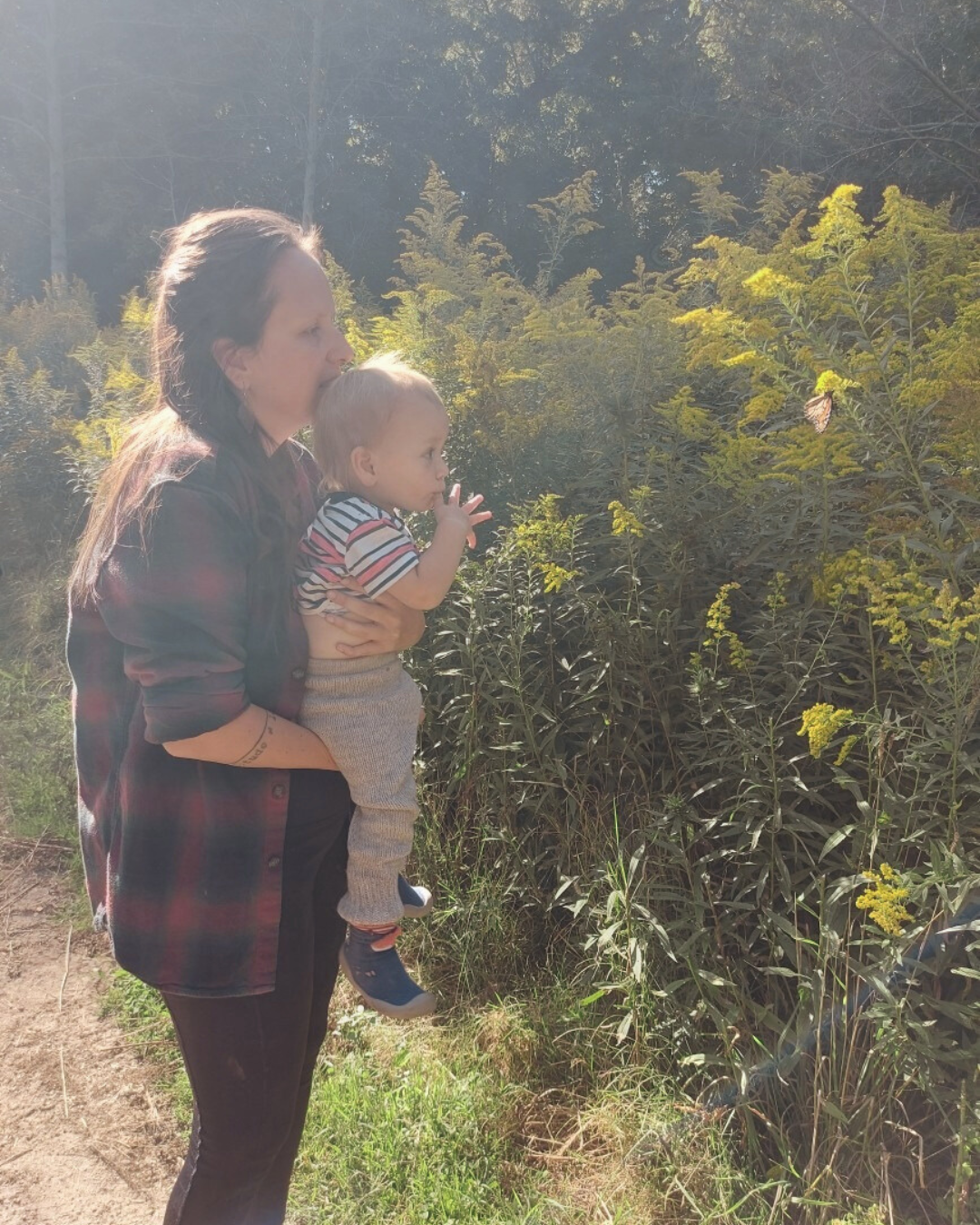 A woman holding a young child while standing outdoors near green foliage and yellow wildflowers on a sunny day.