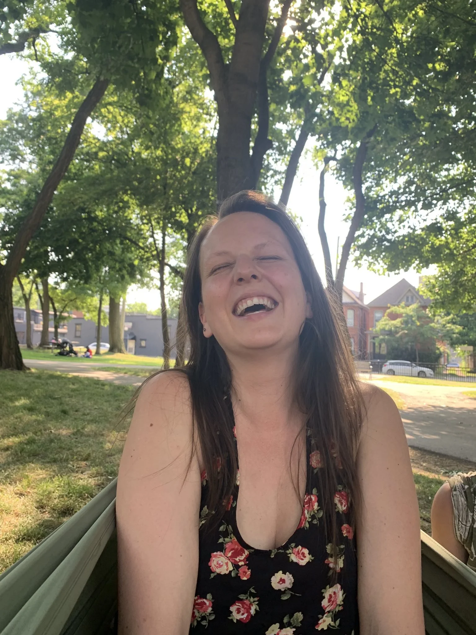 A woman with long brown hair smiling and squinting in a park with green trees, a grass lawn, and a residential neighborhood in the background.