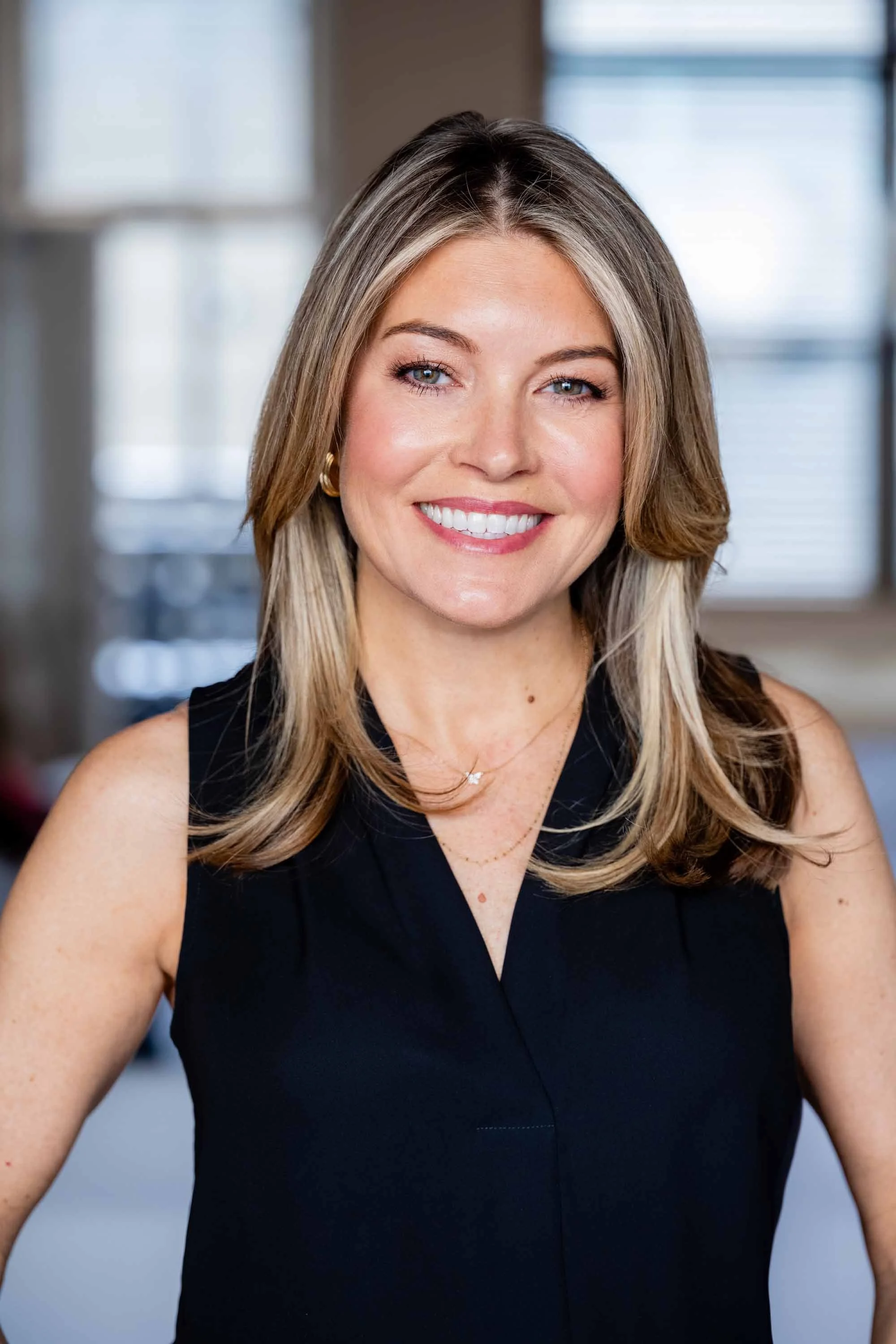 A smiling woman with shoulder-length blonde hair, wearing a sleeveless black top, gold earrings, and layered necklaces, sitting in a brightly lit room with windows in the background.
