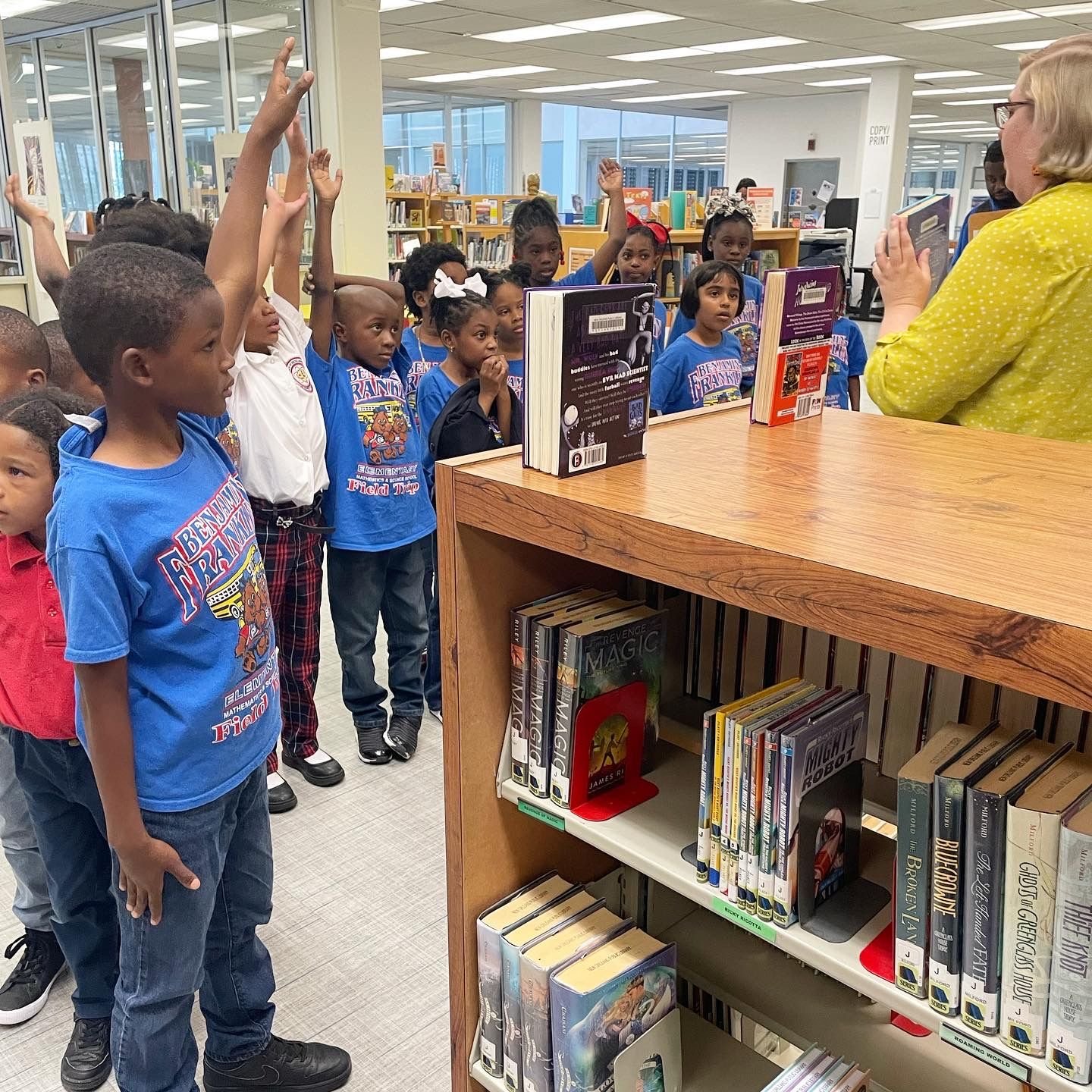 Group of school children raising their hands while listening to a librarian in a library.
