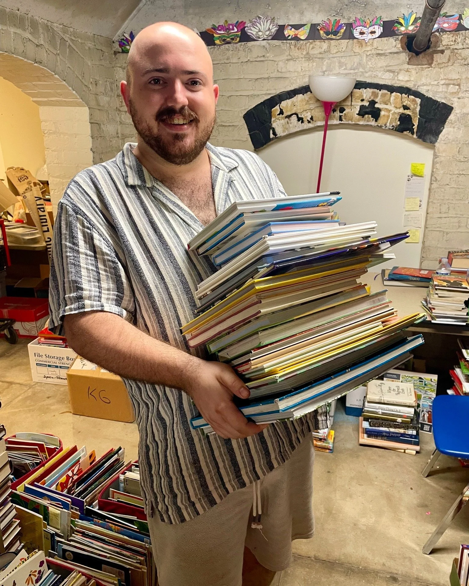 A man with a beard and striped shirt smiling while holding a large stack of files or notebooks in a room with brick walls and various books and supplies around him.