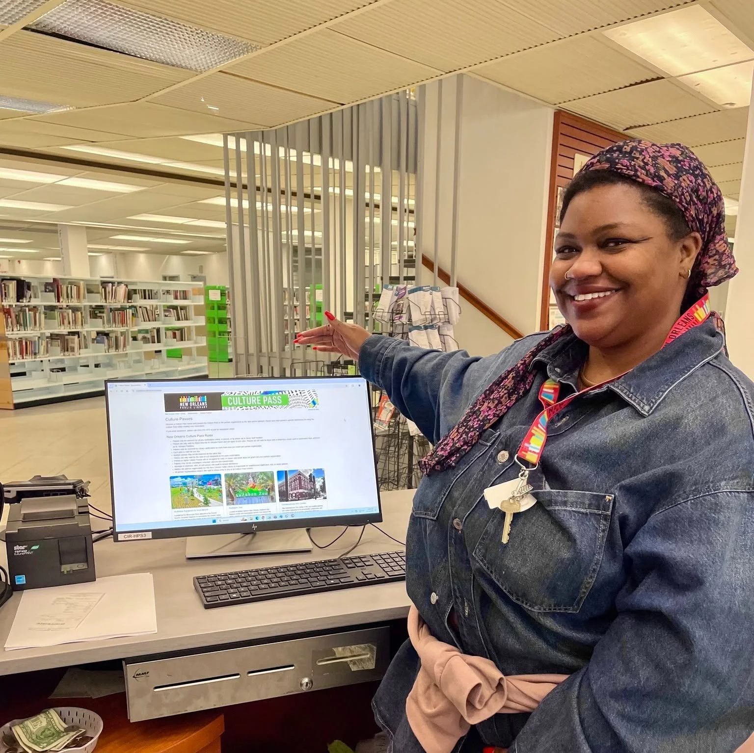 A woman at a computer desk in a library, smiling and pointing to a monitor displaying a website about New Orleans culture passes.