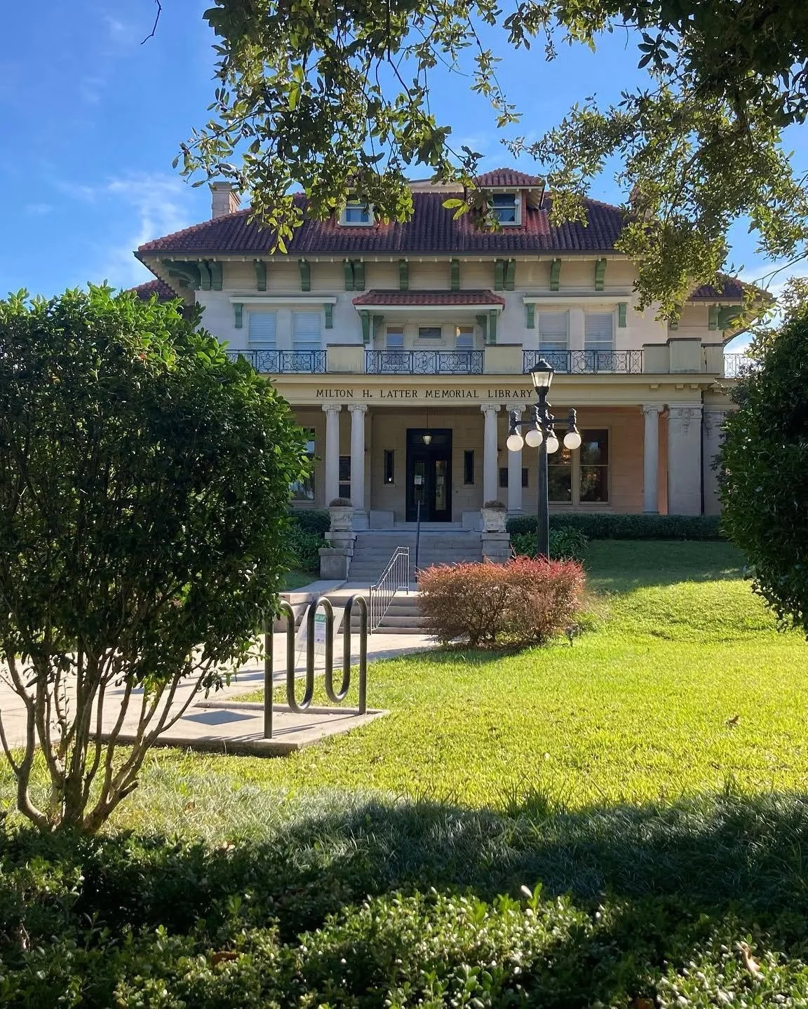 Blue sky and trees overlooking a beige historic building with a red tiled roof, columns, and a sign reading 'Milton H. Latter Memorial Library'.