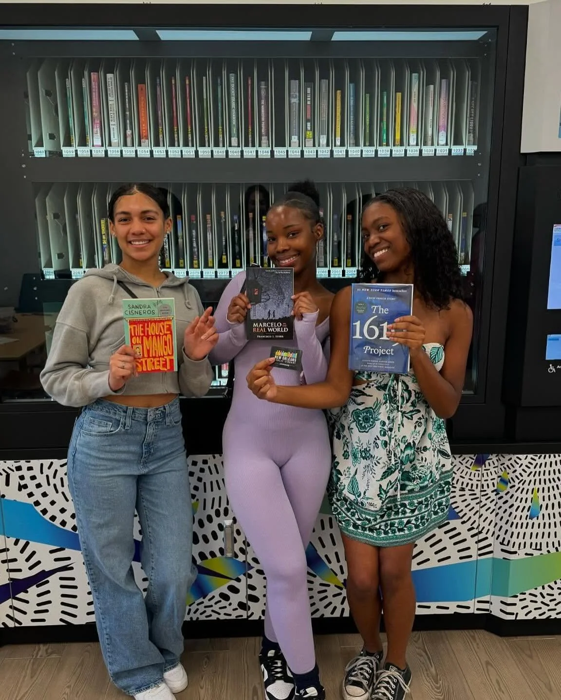 Three young women standing in front of a library book shelf, each holding a book and smiling at the camera