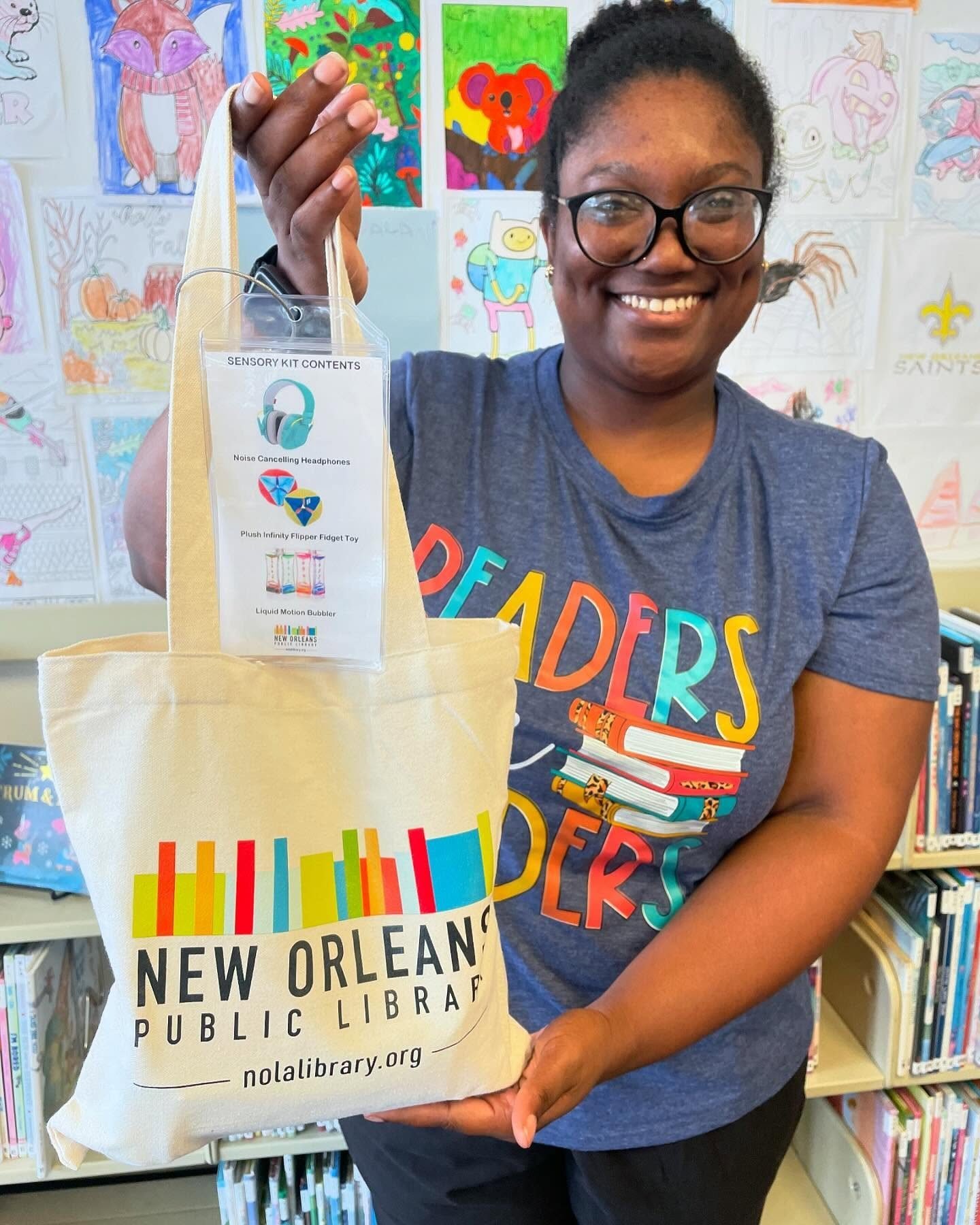 Woman smiling holding a tote bag with New Orleans Public Library logo. Inside the bag is a tag listing sensory kit contents: noise-canceling headphones, plush infinity flipper fidget toy, liquid motion bubbler. Background features colorful children's artwork and a bookshelf.
